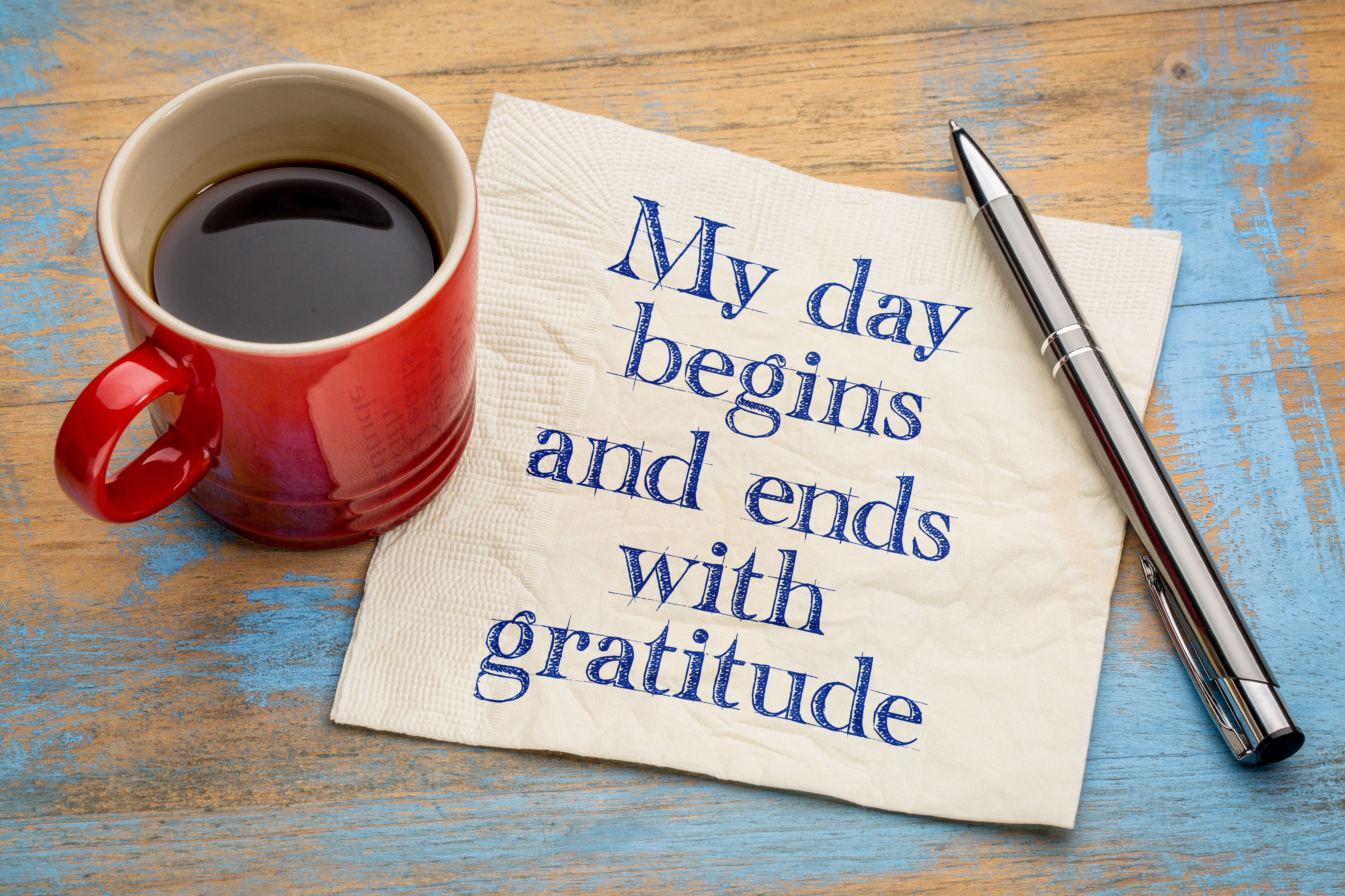 An image of a full coffee cup sat on a blue-washed wooden surface beside a napkin with a positive affirmation reading My day begins and ends with gratitude written on it