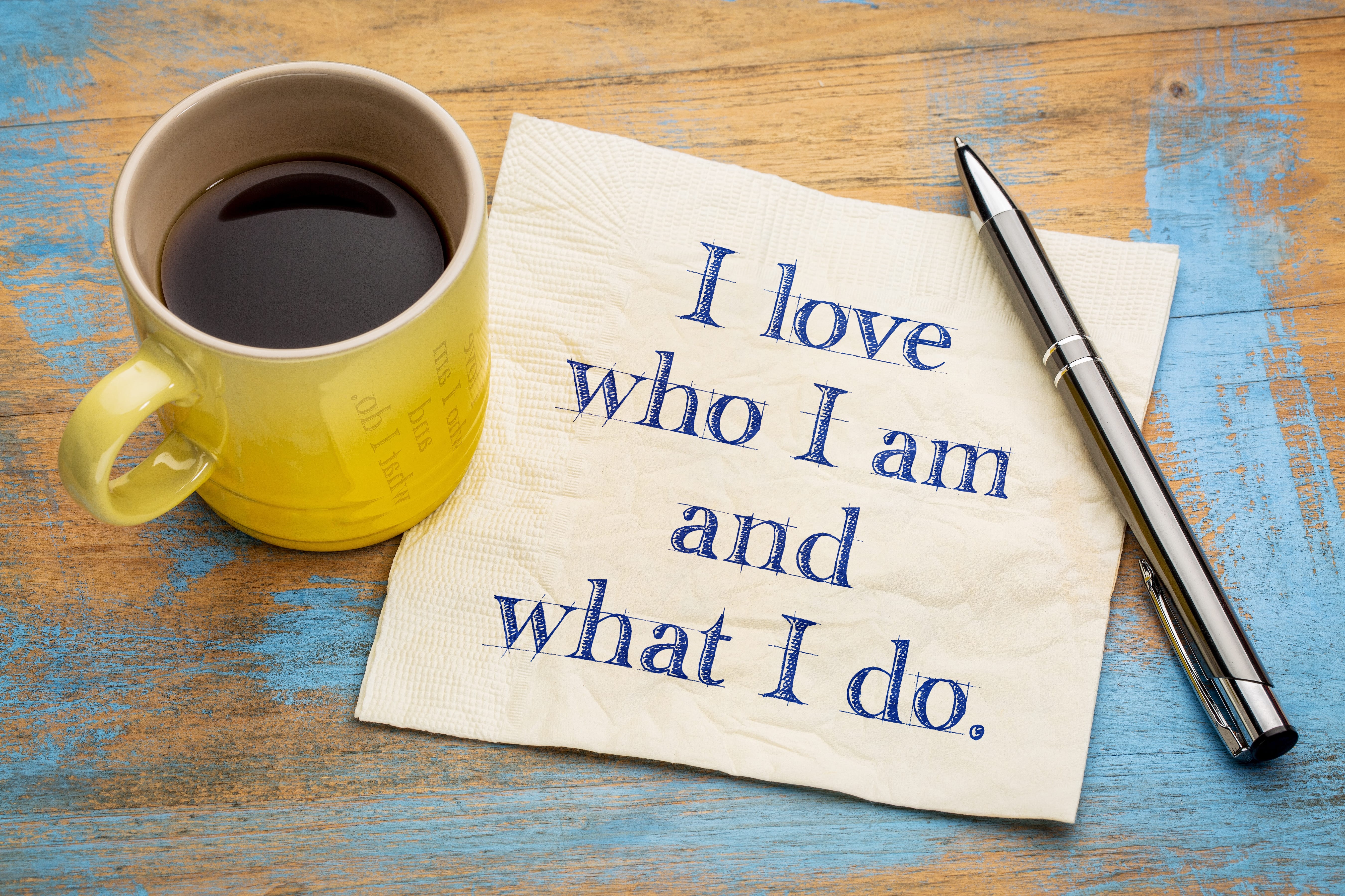 A yellow coffee cup on a blue-wash wooden surface beside a napkin with the positive affirmation I love who I am and what I do written on it.