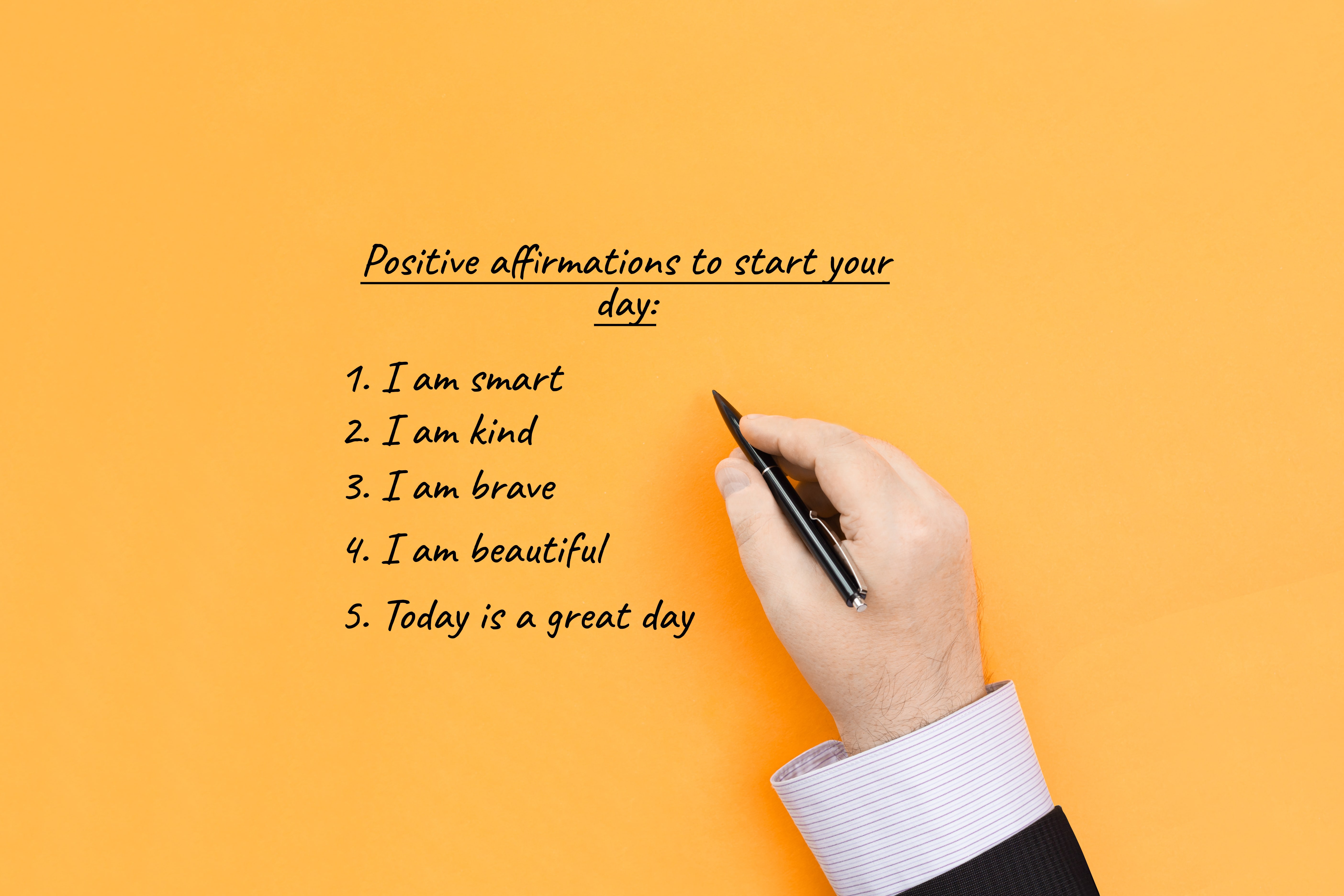 A hand holding a pen writing a list of positive affirmations to start your day.