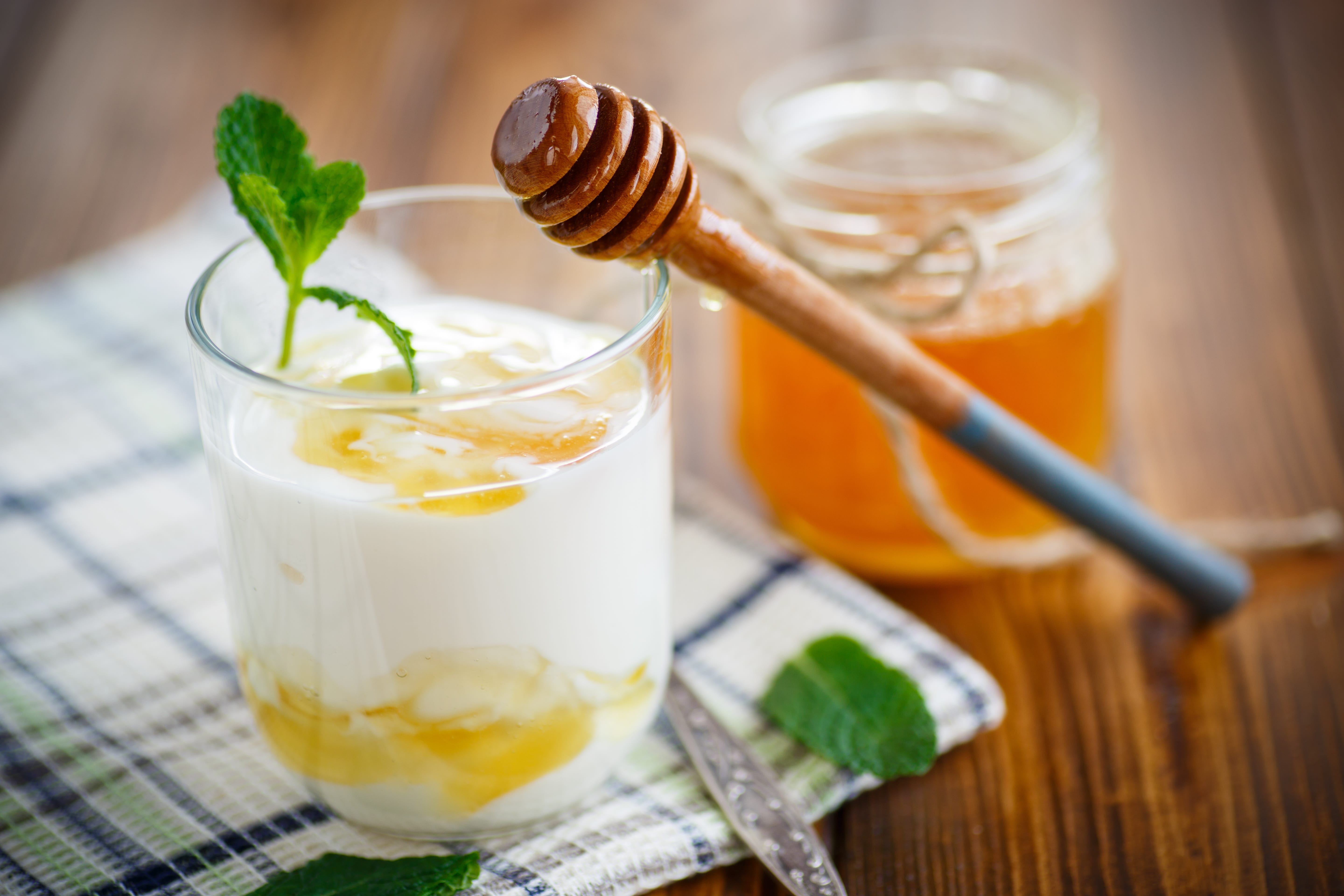 Yogurt and drizzle of honey in a glass with a sprig of mint. A wooden honey drizzler leans against the glass and a pot of honey is in the background.