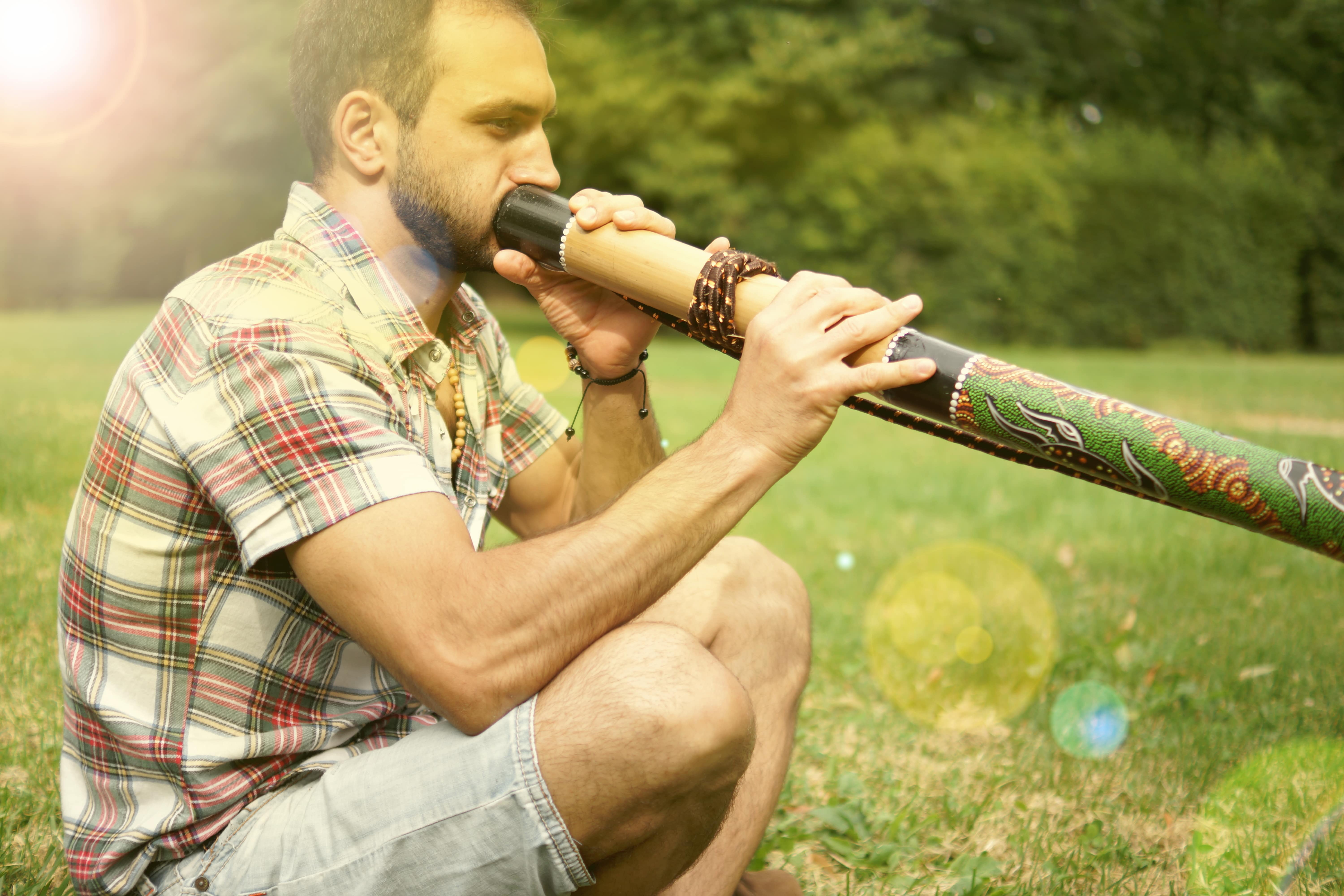 A man sitting in a field on a sunny day blowing into a didgeridoo.