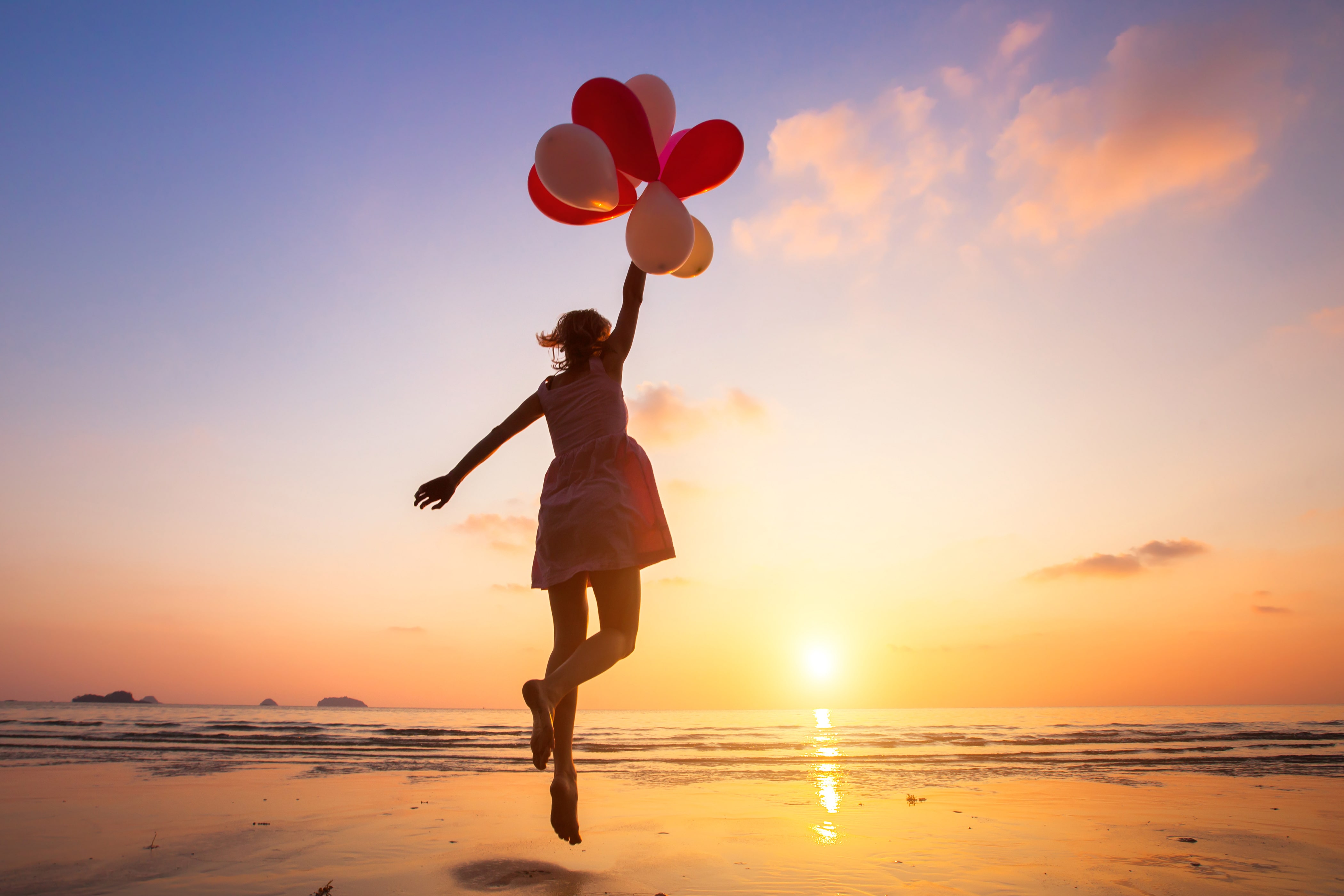 Image of a lady holding a handful of balloons as she floats above the sandy coastal beach at sunset.