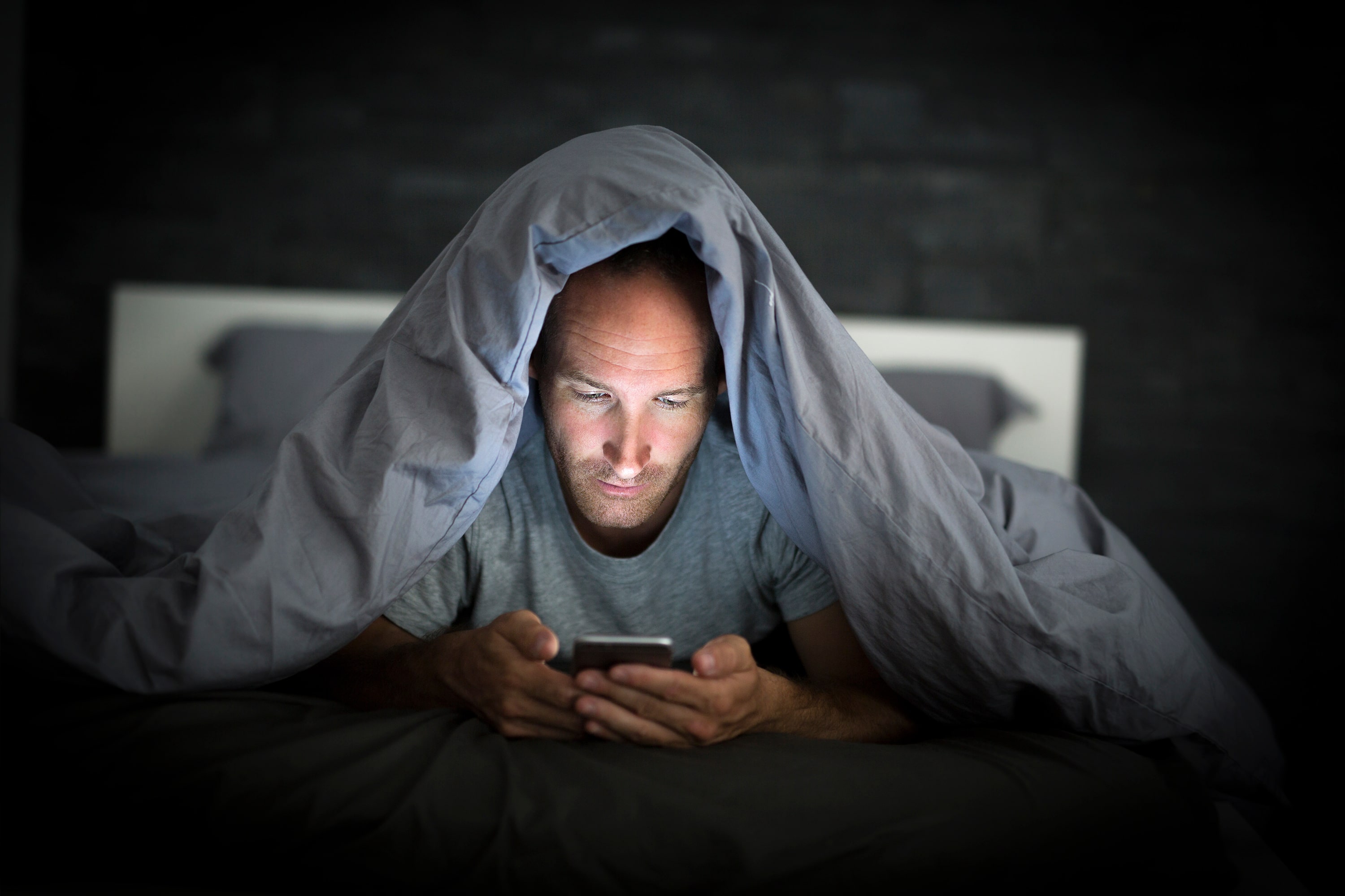 A man burrowed under his duvet in bed while scrolling through social media on his smartphone.