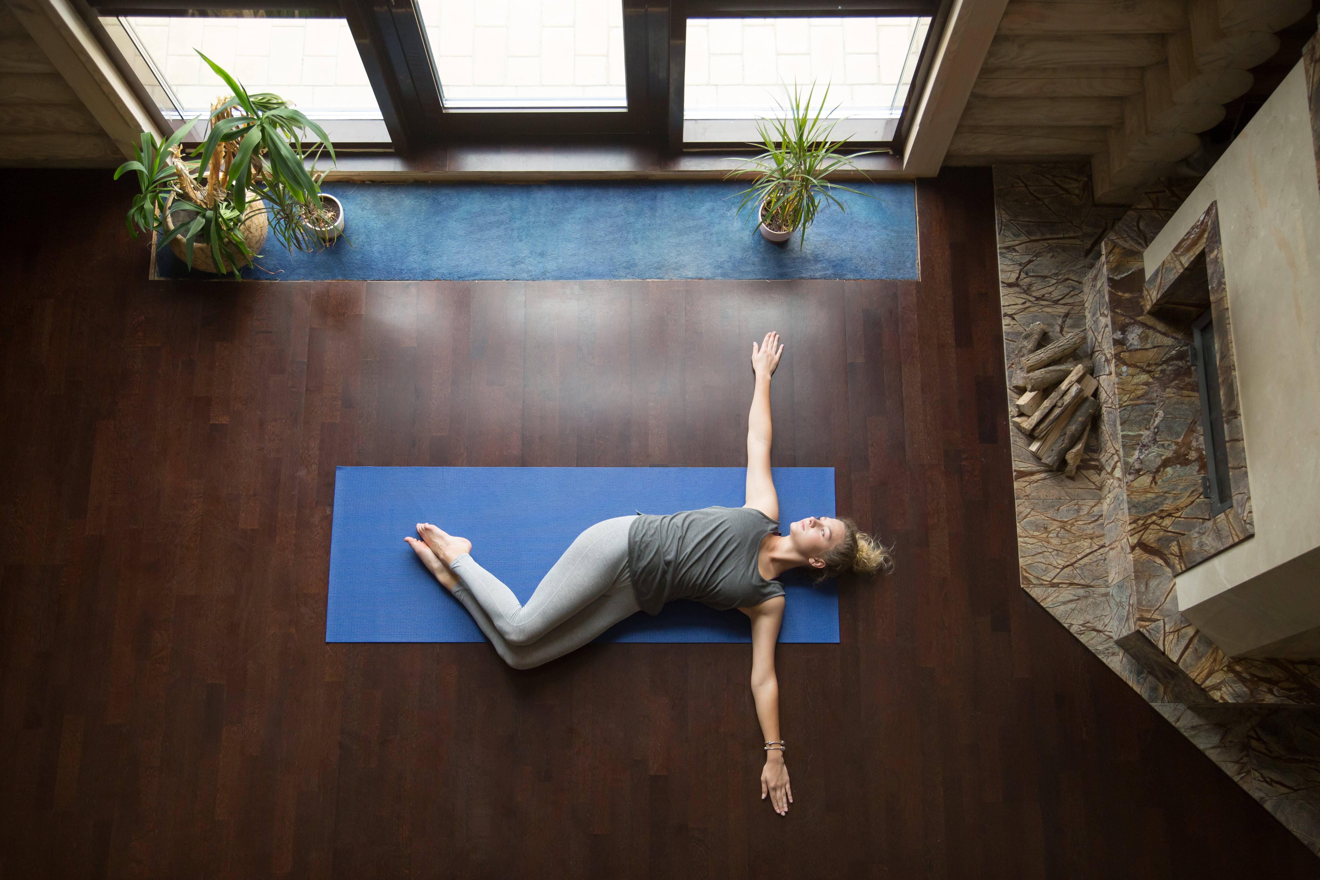 A lady practicing the supine twist yoga pose. The photo is taken from an aerial view and a stone fireplace and several house plants enhance the calming visual