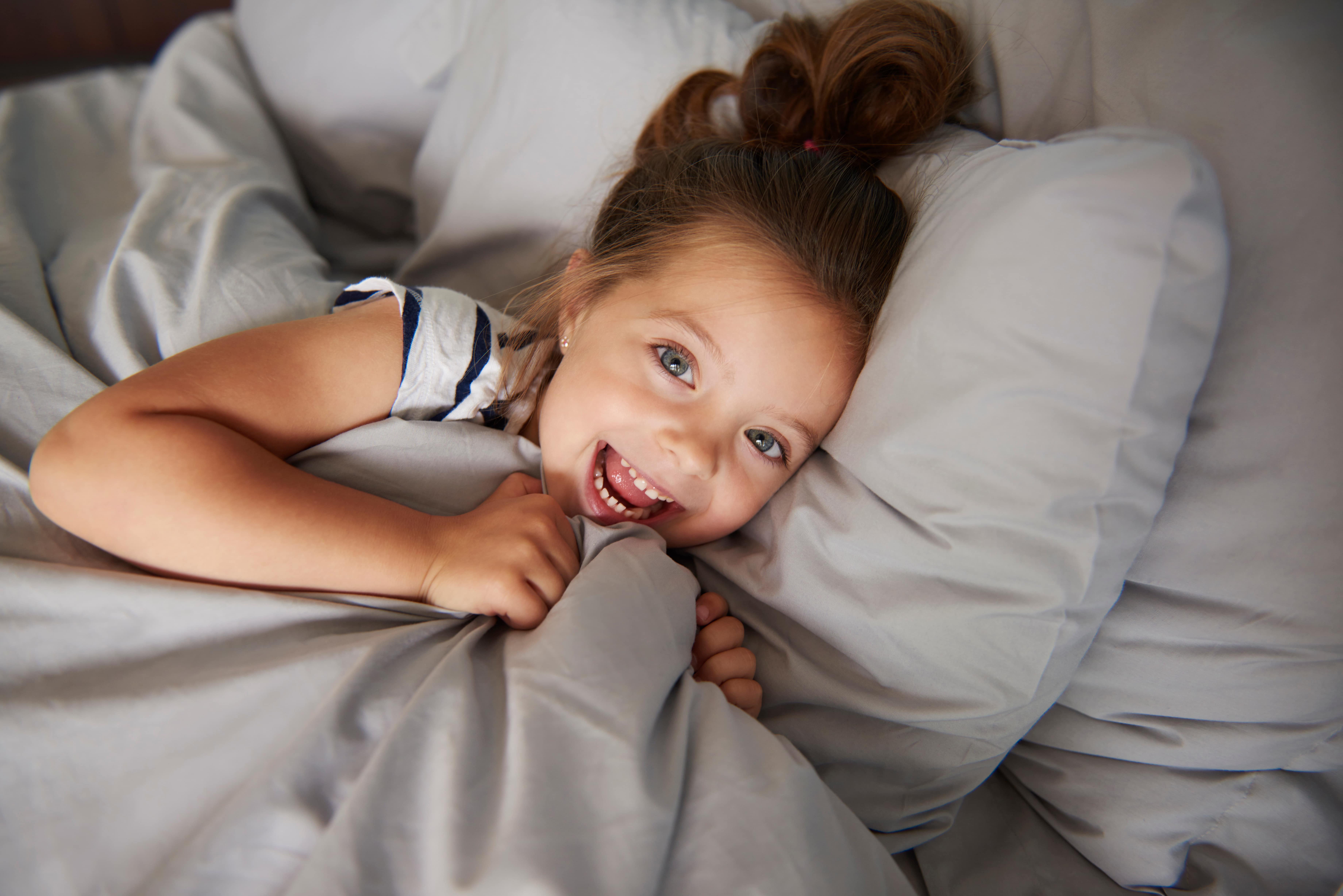 Girl in bed smiling and clutching duvet under her chin.