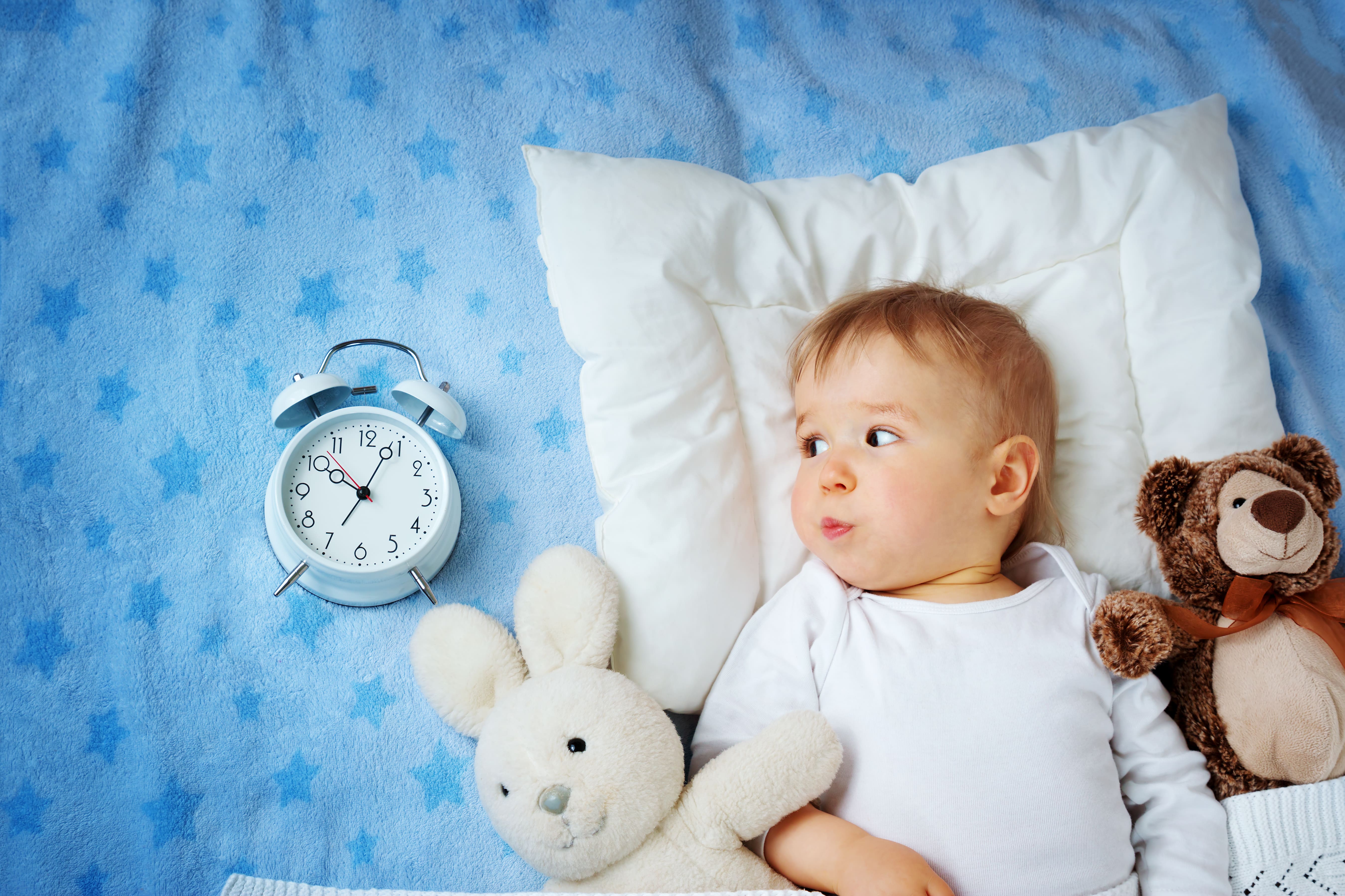 Toddler looking at an alarm clock to their side and lying on pillow and starrry blanket flanked by teddies either side.