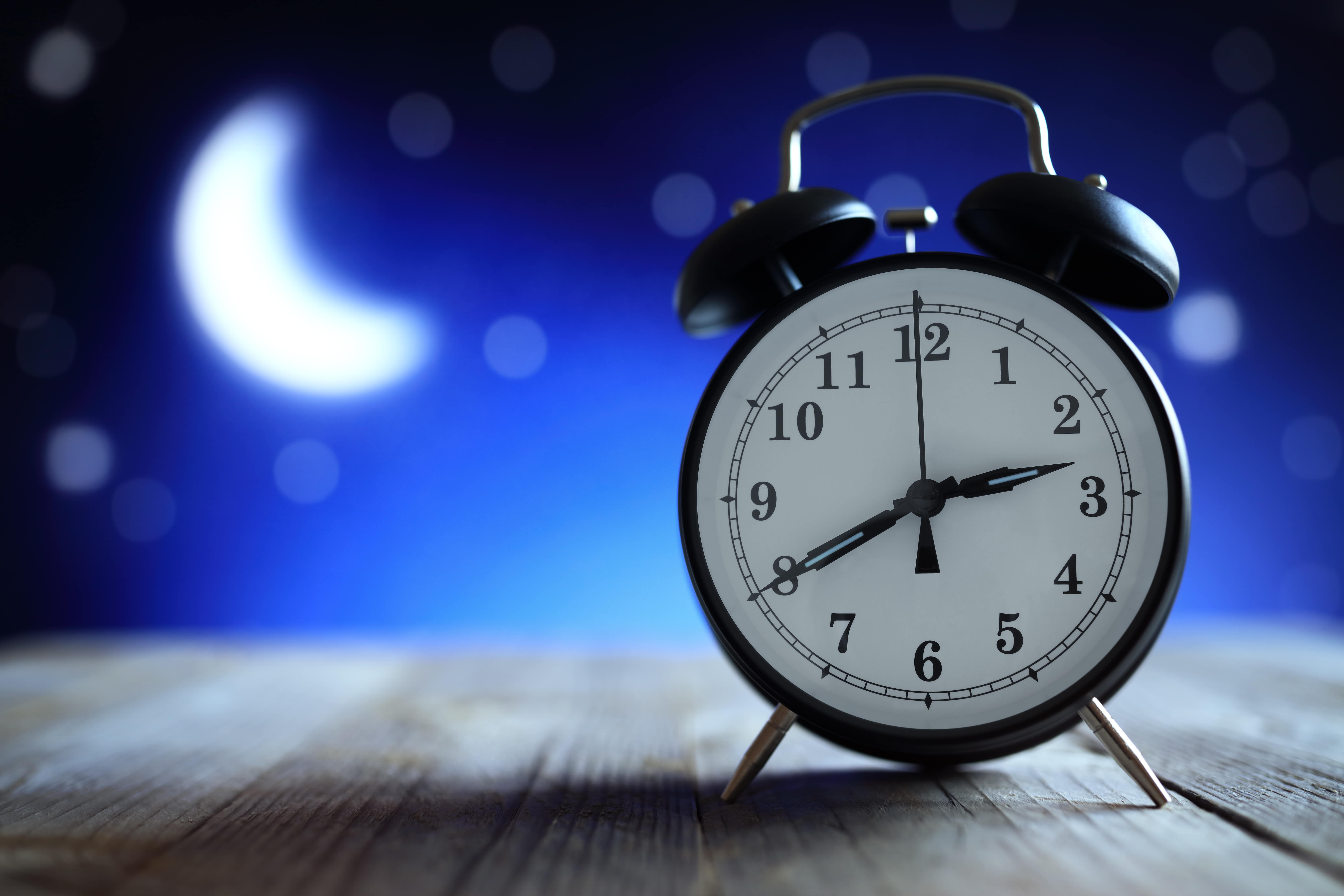 A traditional alarm clock is in the foreground on a wooden floor. The background is blue with a blurred image of a white crescent moon.