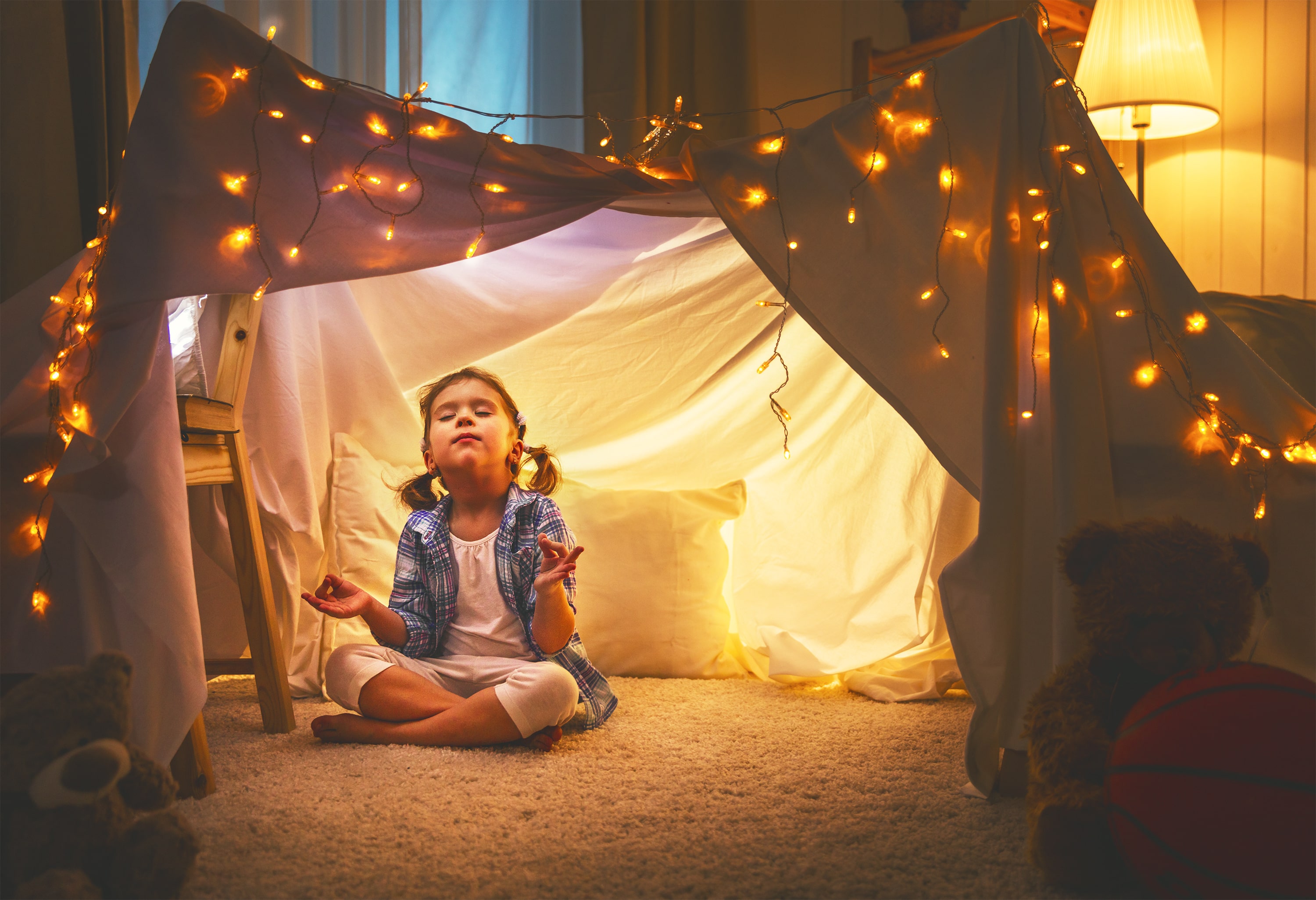 A little girl sat in a home-made den adorned with fairy lights while doing some bedtime yoga