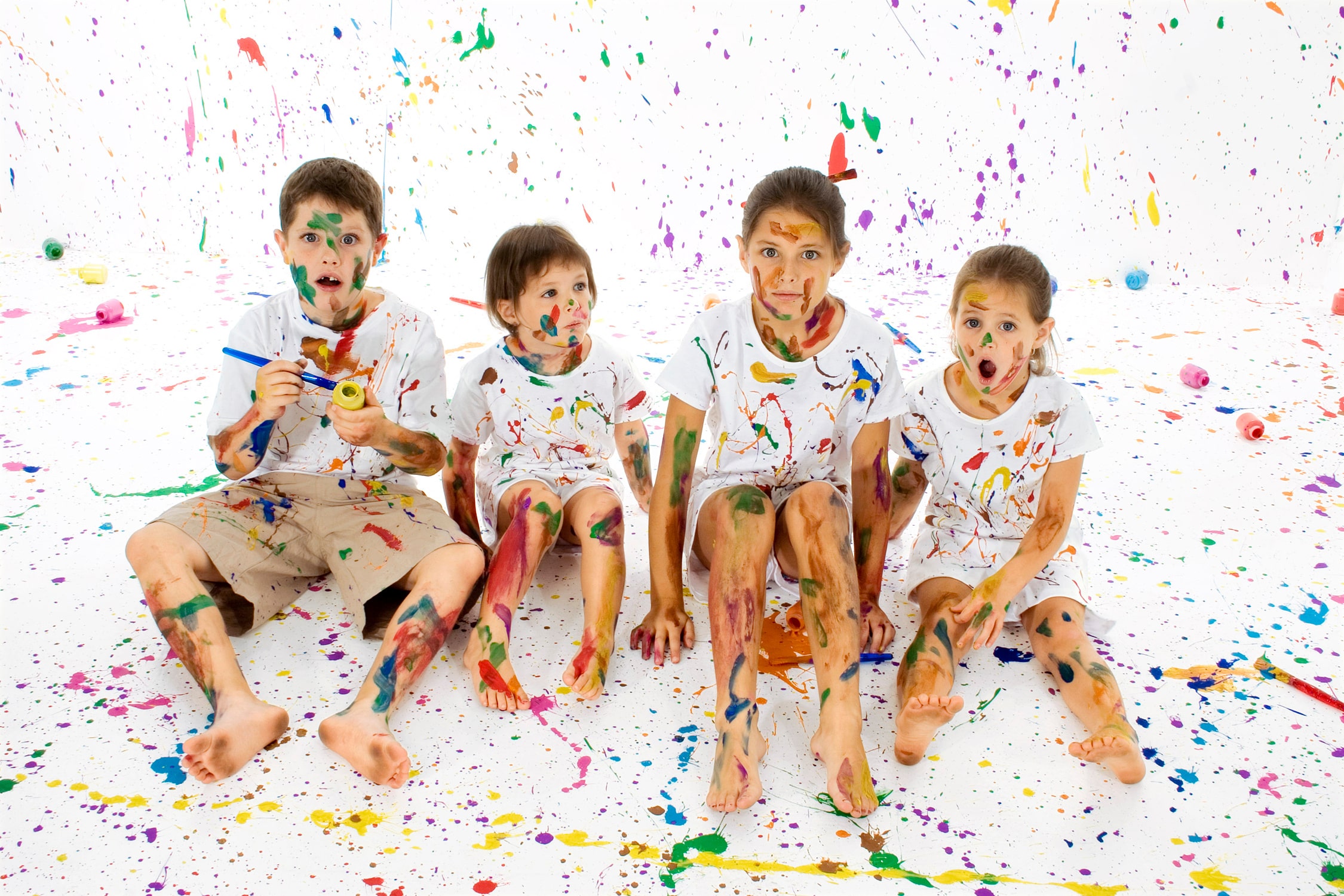 four children sit in a white studio. They are covered in splashes of paint as is the wall an floor of the studio.