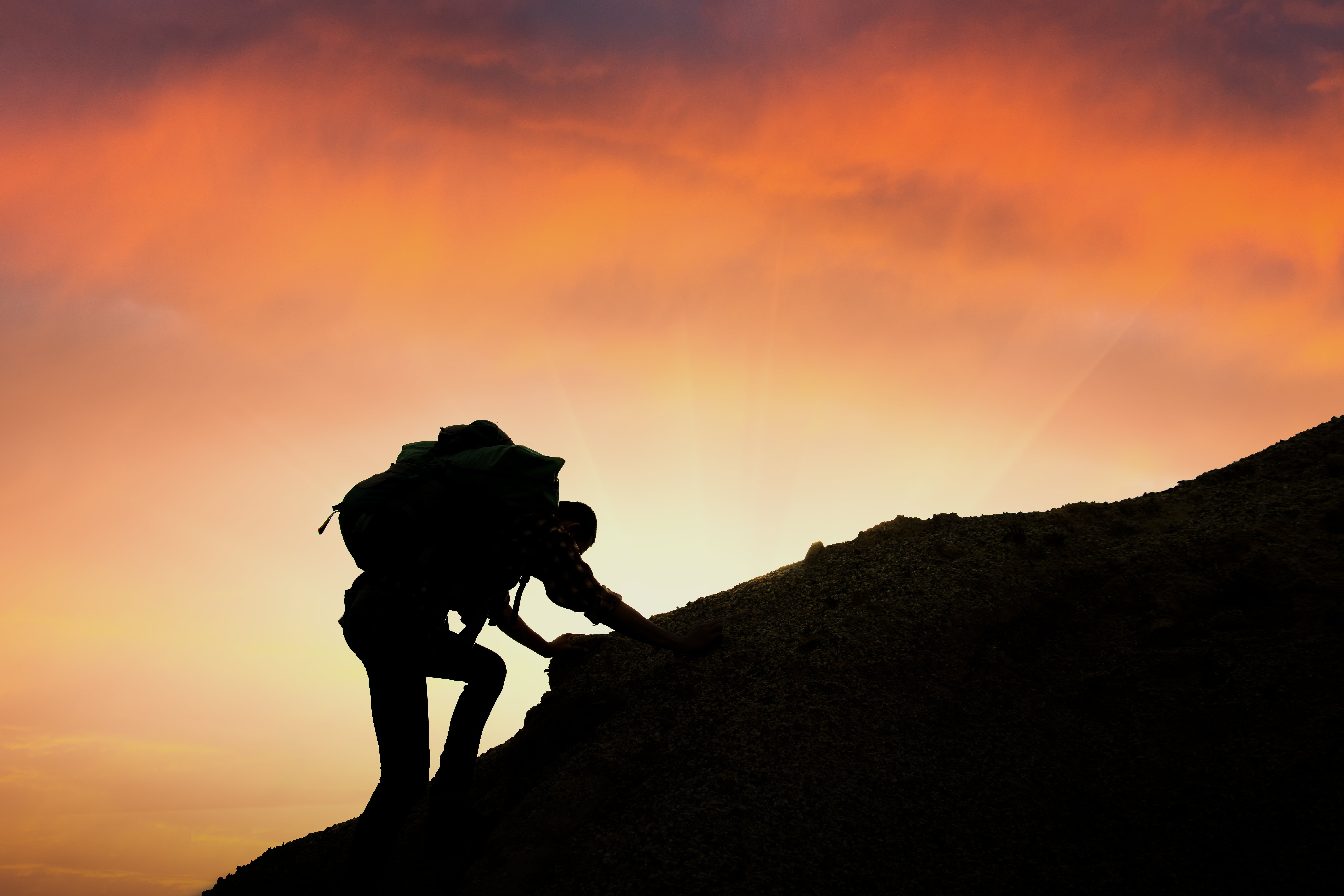 A person climbing a mountain with a large rucksack on their back silhouetted against the sunset