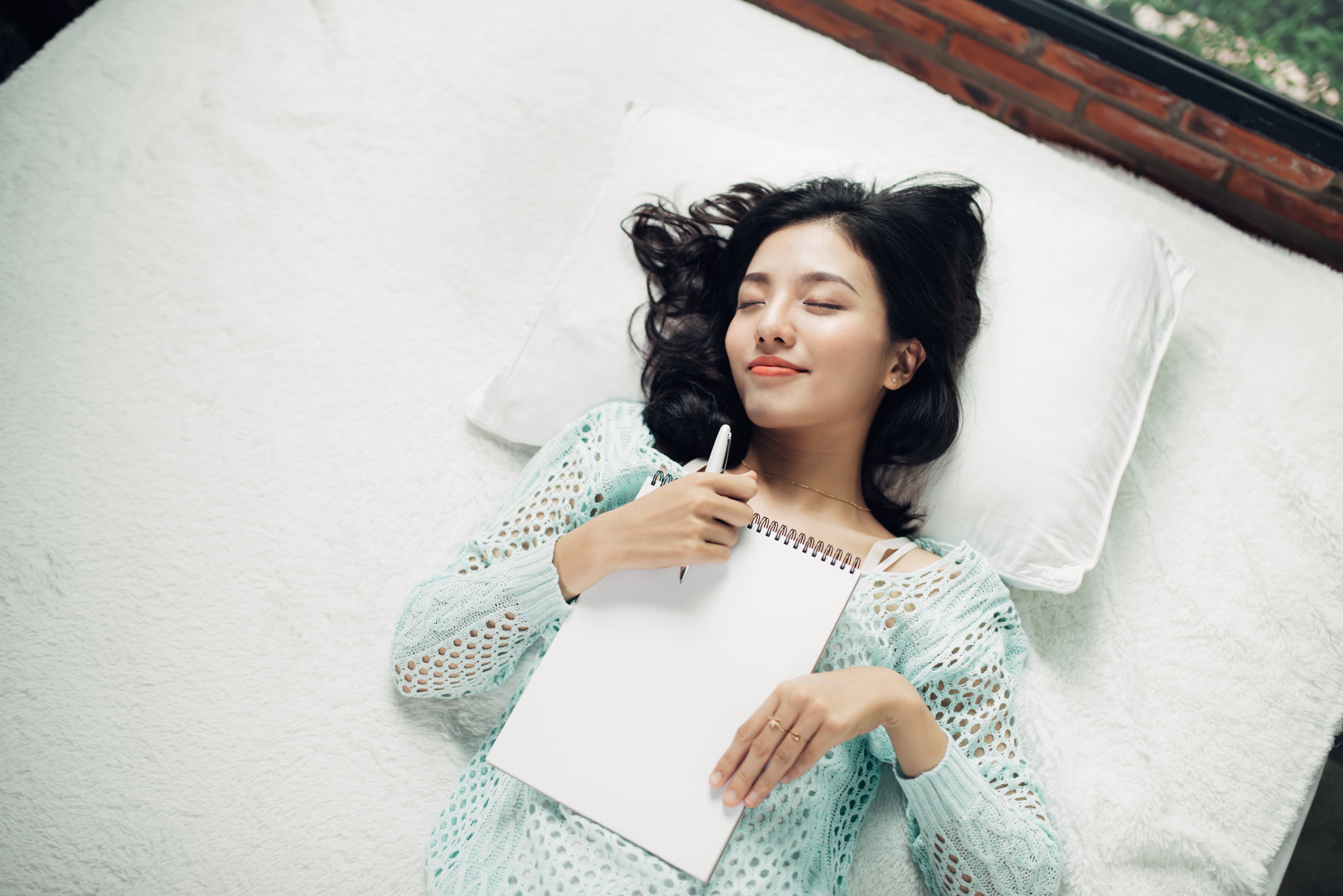 Woman lying back on a bed with her eyes closed and smiling. She clasps a pen in one hand and a notepad rests on her chest.