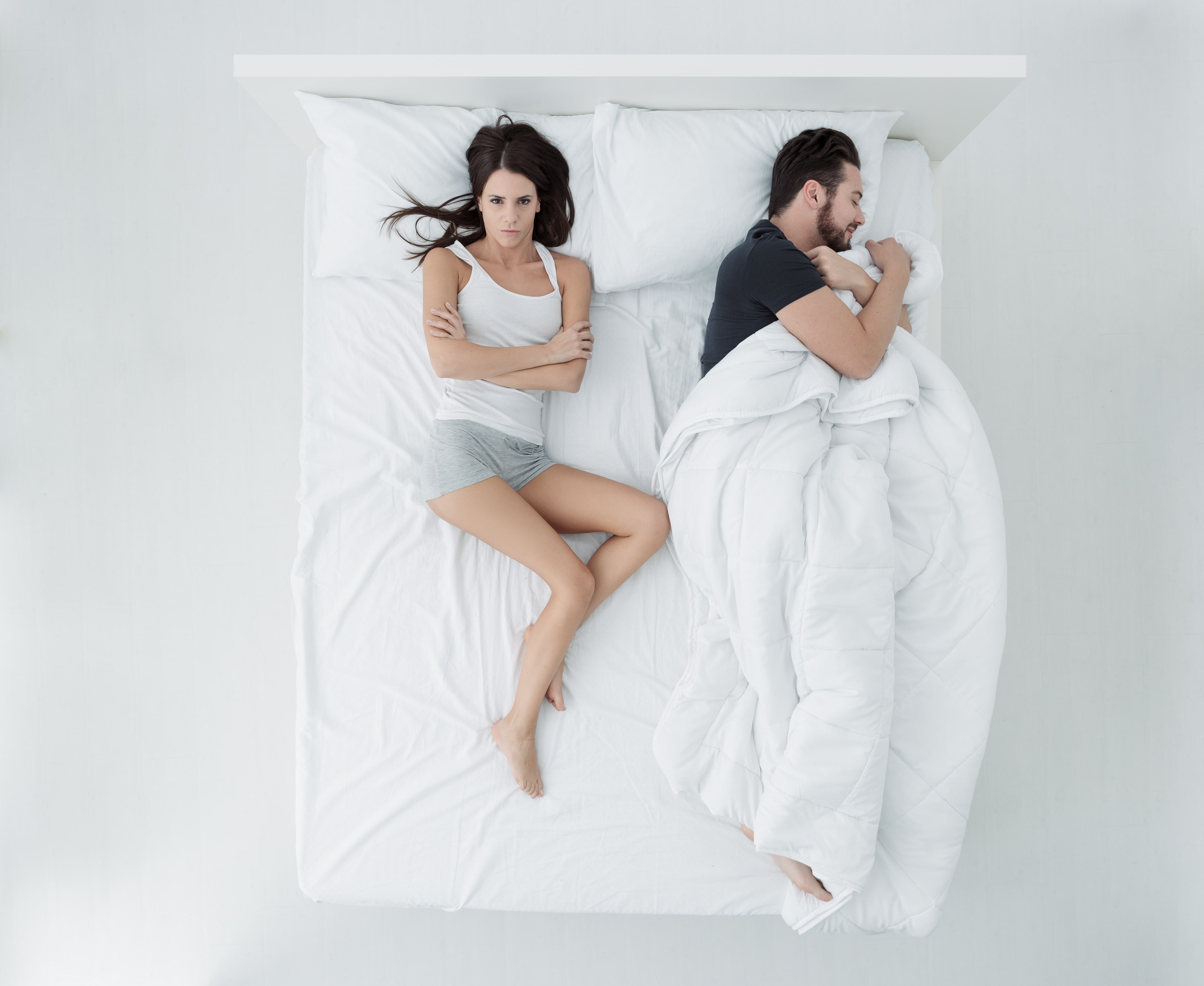 Man and woman in bed. Man is sleeping wrapped in a duvet and woman looks angry as she isn't covered by any of the duvet.