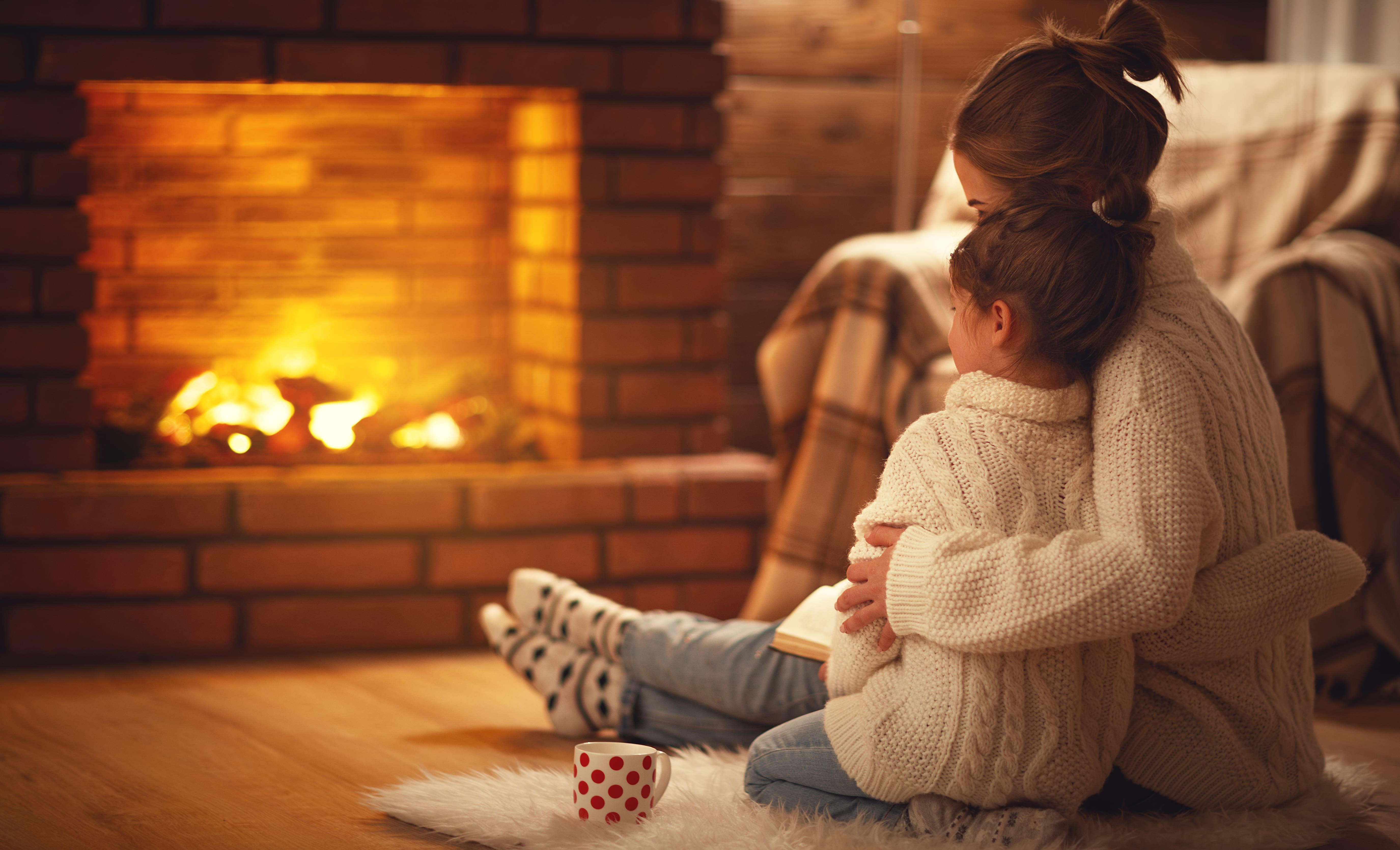 A mother and child, snuggling in the warm glow of the fire. Both are wearing cable knit jumpers. 