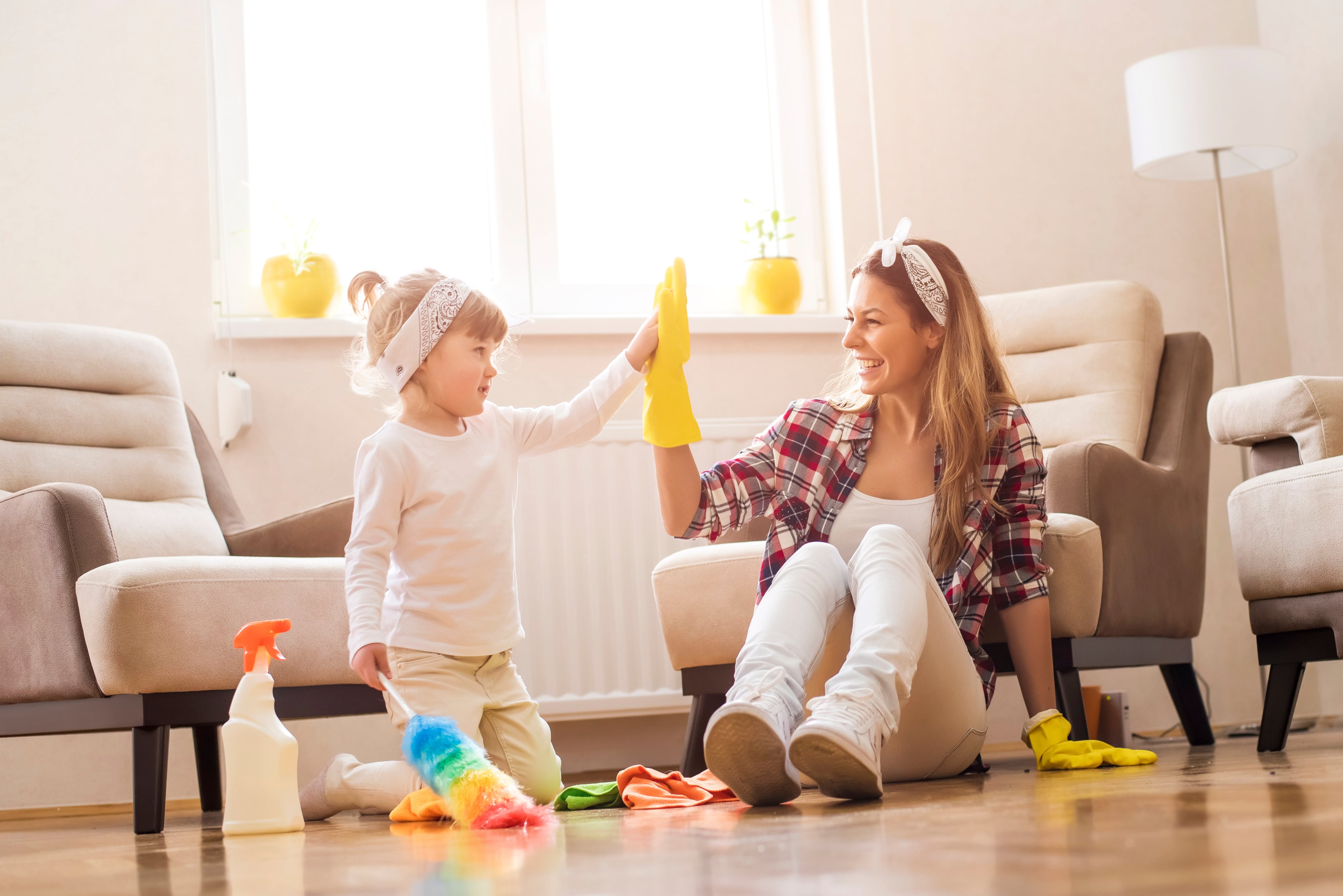 A mum and daughter high-fiving after a successful spring cleaning session. The mum is smiling enthusiastically and wearing marigolds, the daughter holds a multi-coloured feather duster in her other hand.