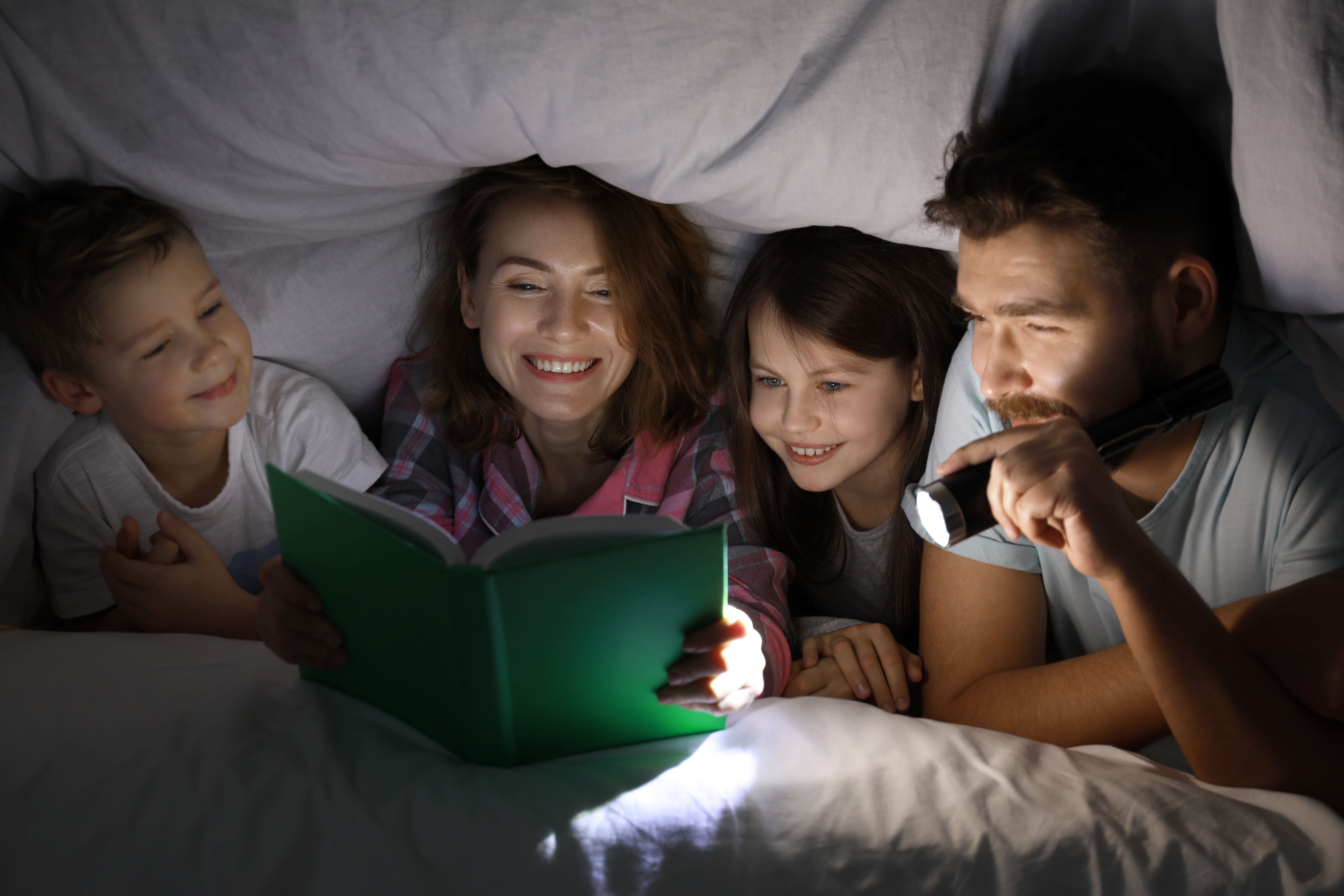 A family of four huddled under a duvet while reading bedtime stories. Mum is reading the book while dad holds a torch to that everyone can see.