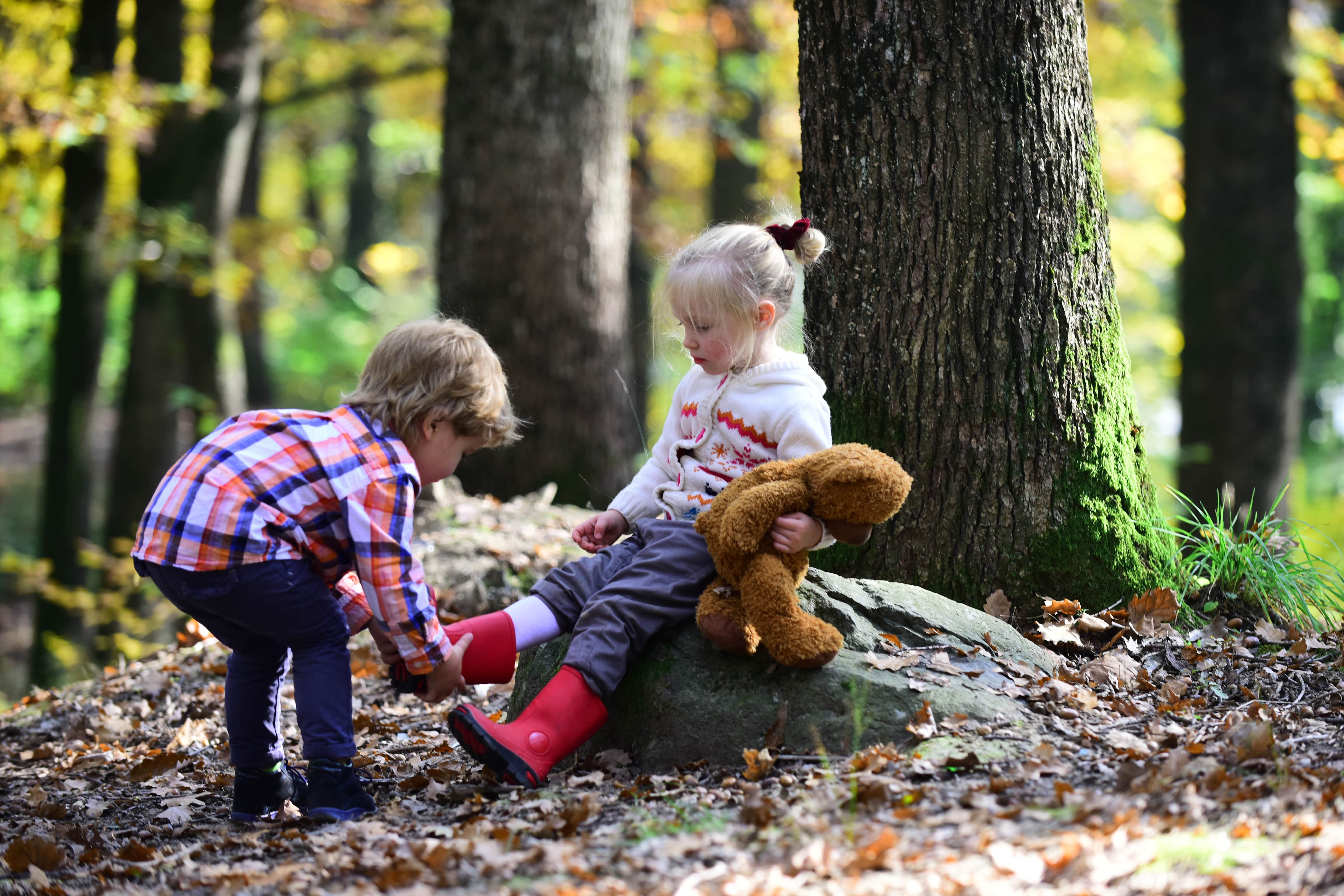 A little boy is helping a little girl take off her wellies. They are in a wood.
