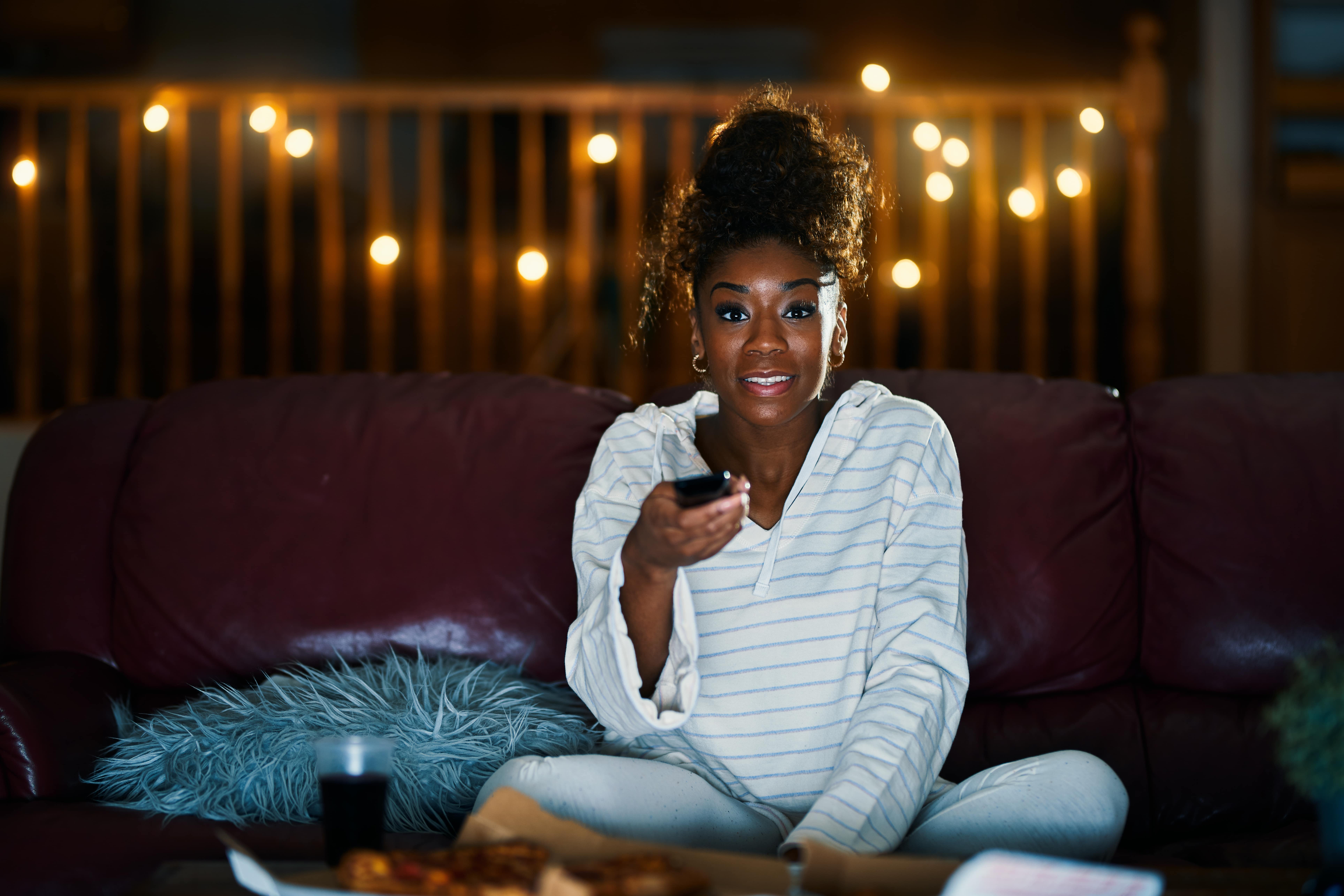 Smiling woman pointing a television remote towards the foreground and seems to be watching T.V. It is dark and there are fairy lights strung up in the background.