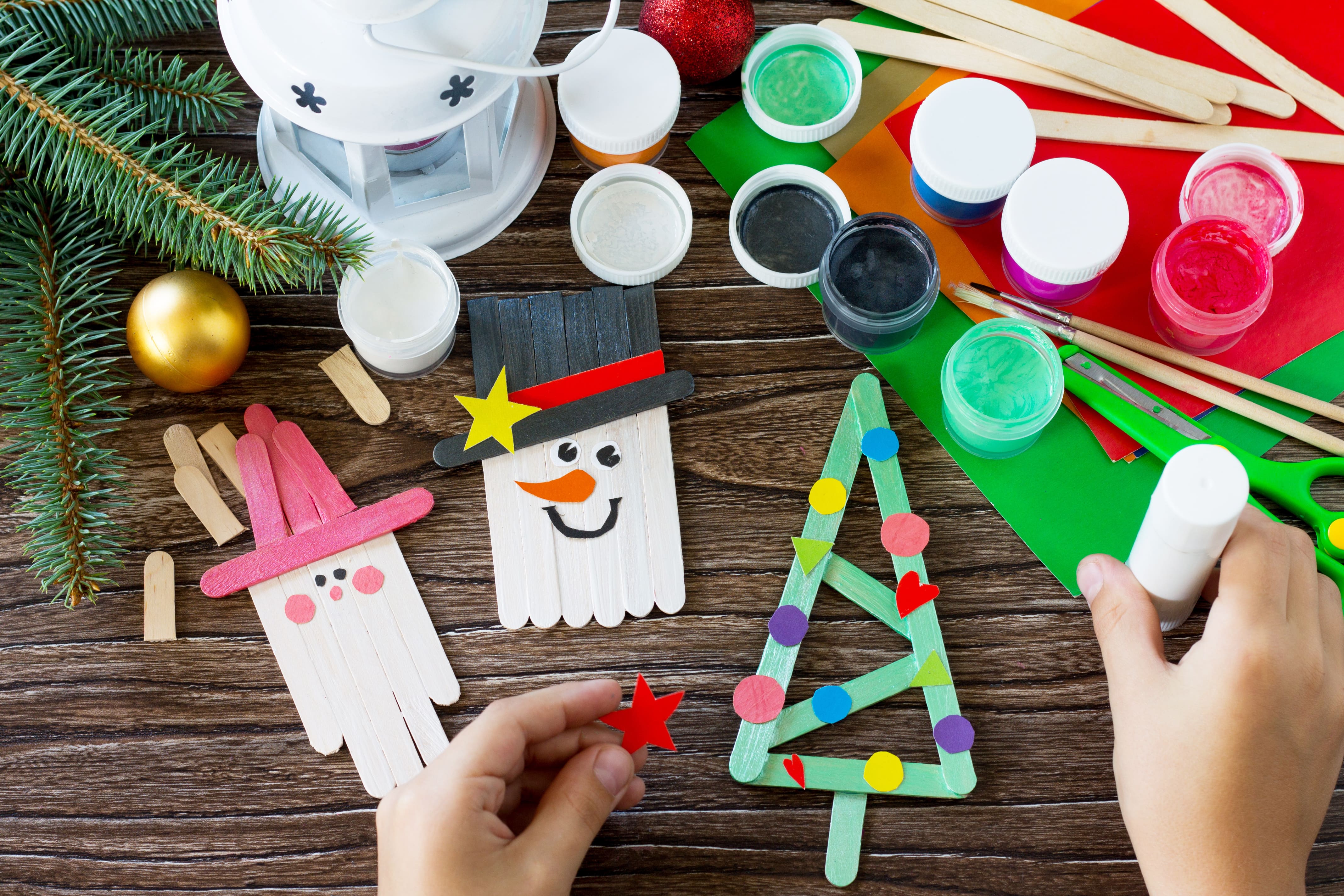 Festive Christmas crafting in session. Open pots of paint, glue, and wooden lolly sticks lie against a backdrop of a wooden table top and some coloured card.