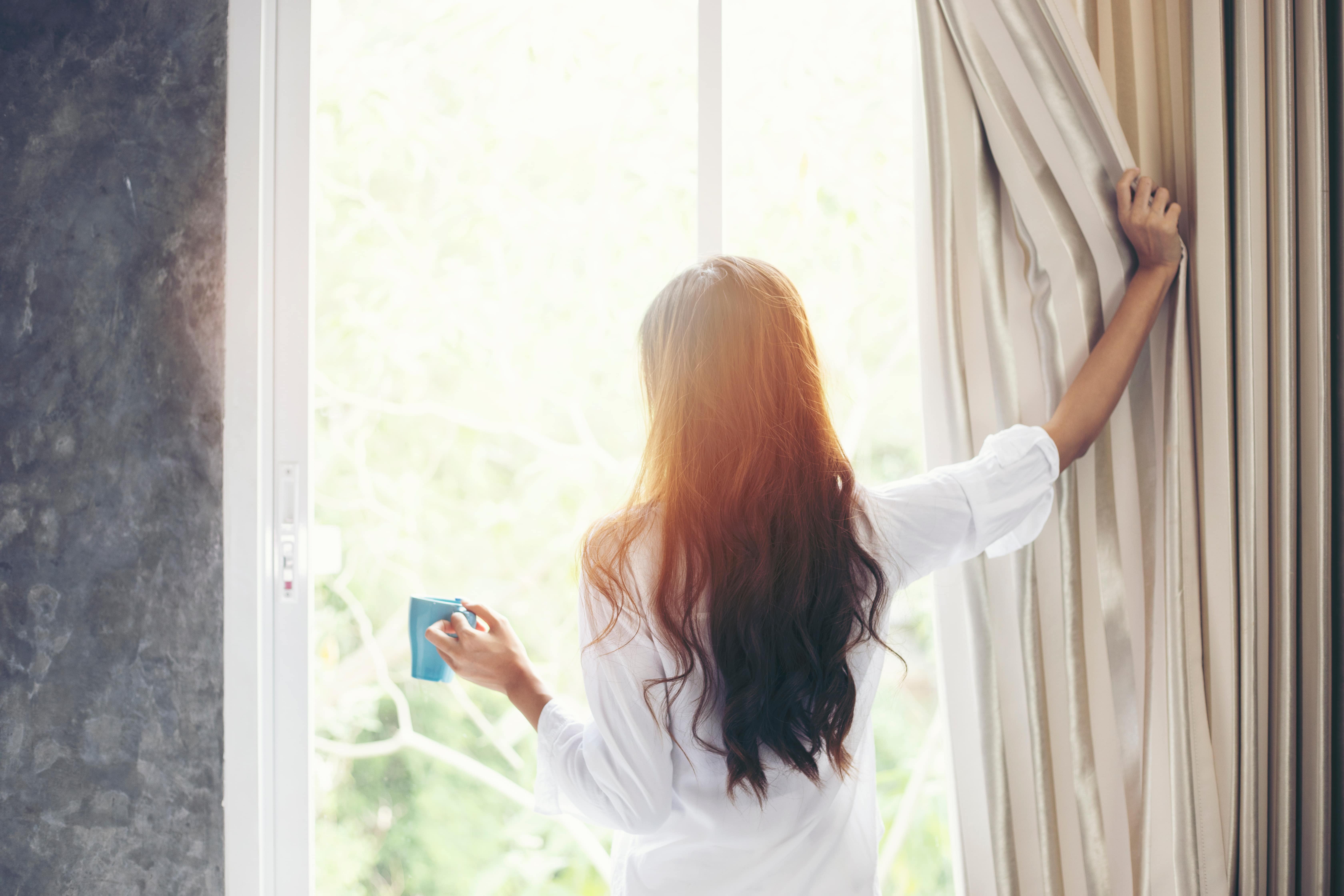Woman opening the curtains on patio doors to bright sunshine and holding a mug.