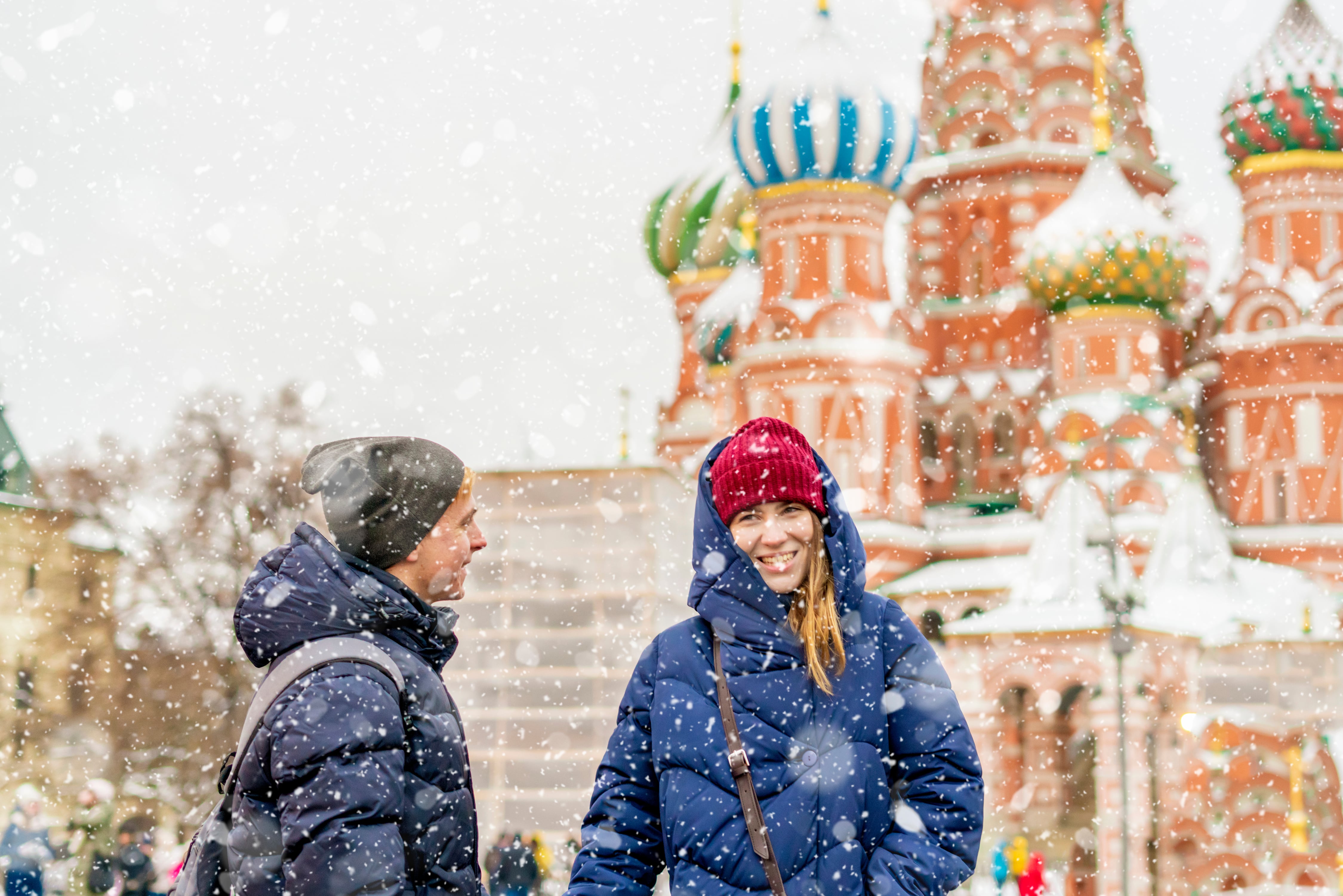 Man and woman wearing winter clothing of jackets and hats stand smiling outside the Kremlin in Moscow. It is snowing.