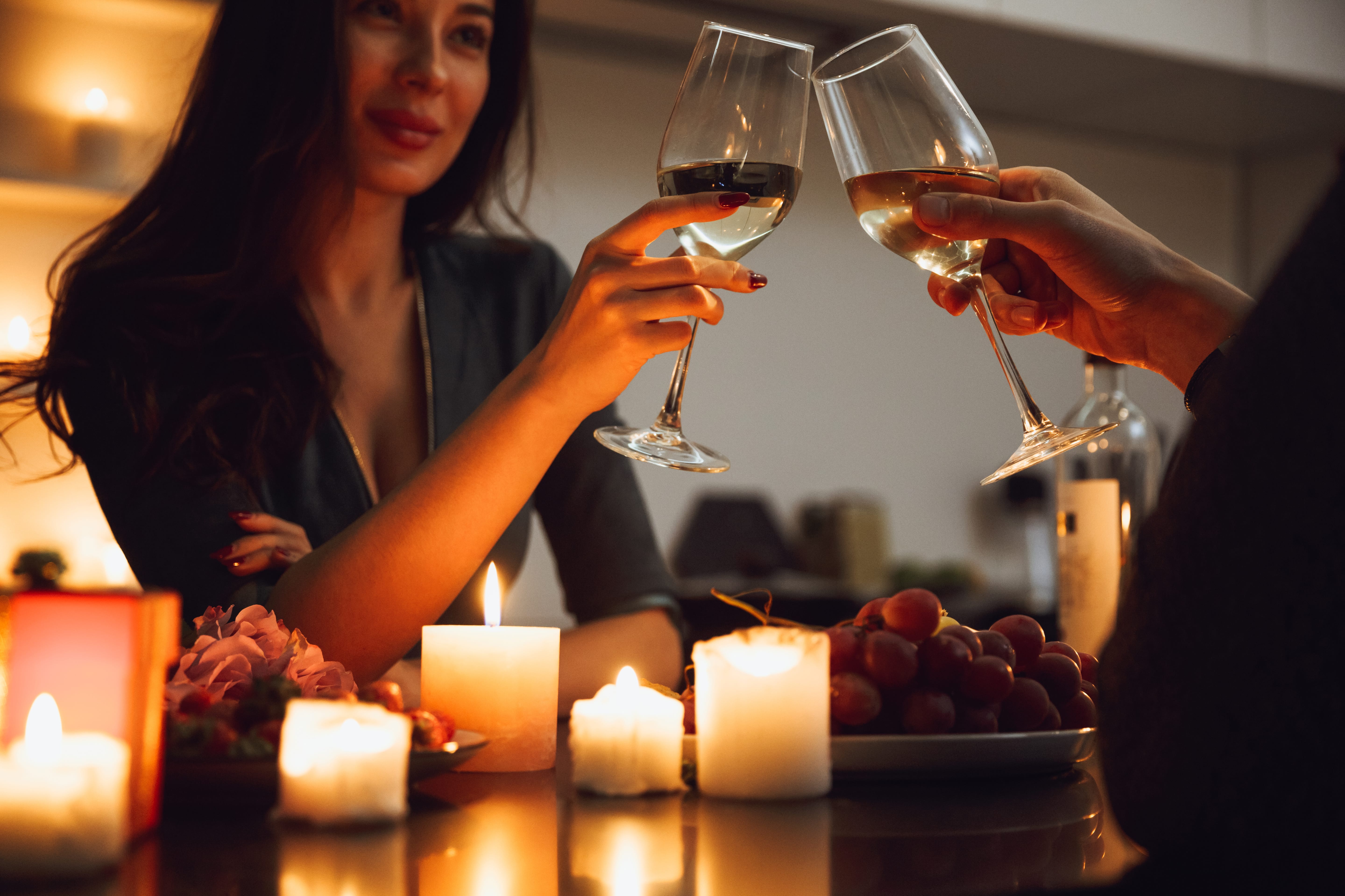 A couple clinking wind glasses by candlelight on a date. A bowl of grapes sits on the table top and is surrounded by multiple candles