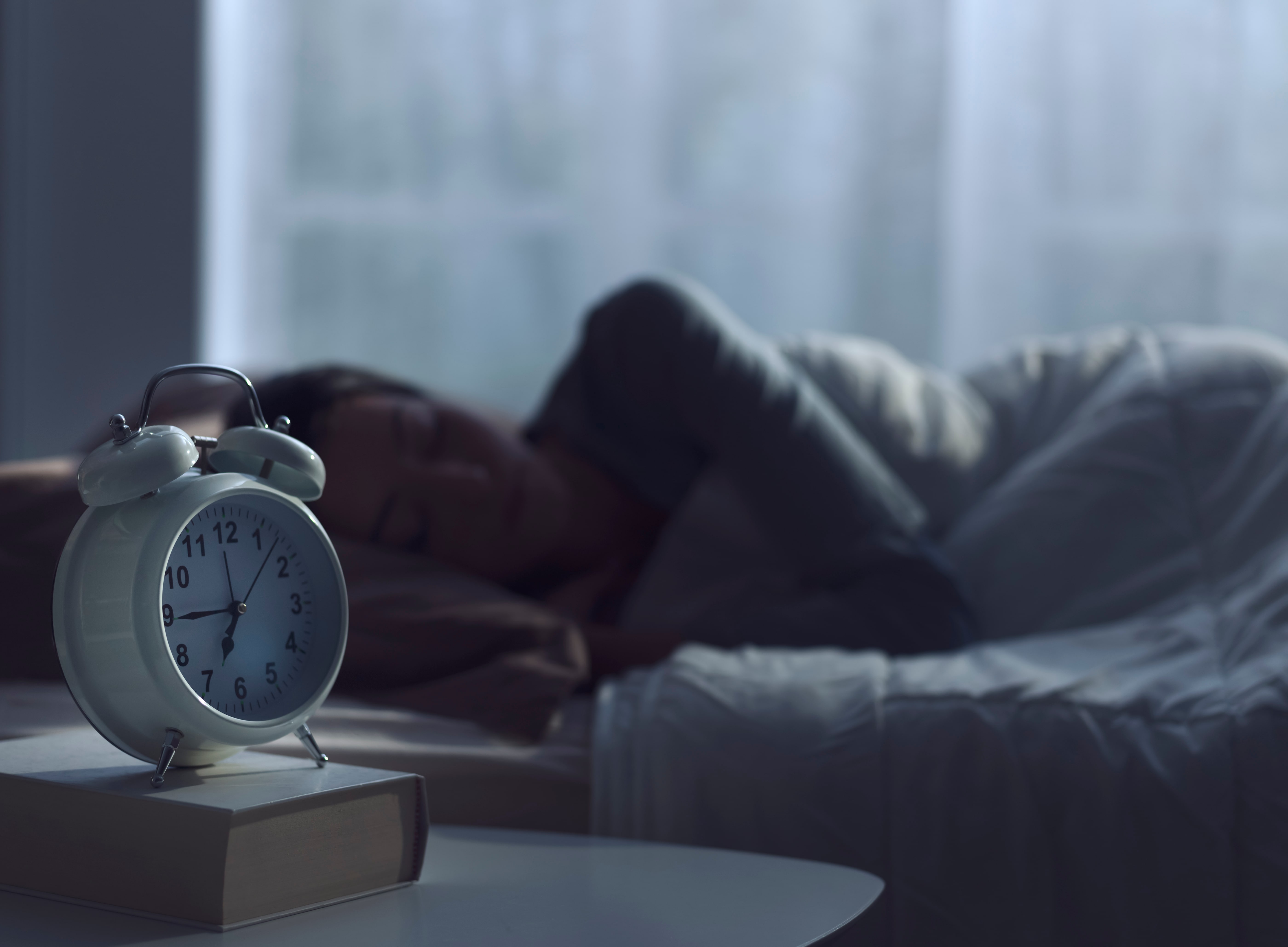 A lady asleep in bed. A retro alarm clock displaying the time 6:45 am is in the foreground of the image