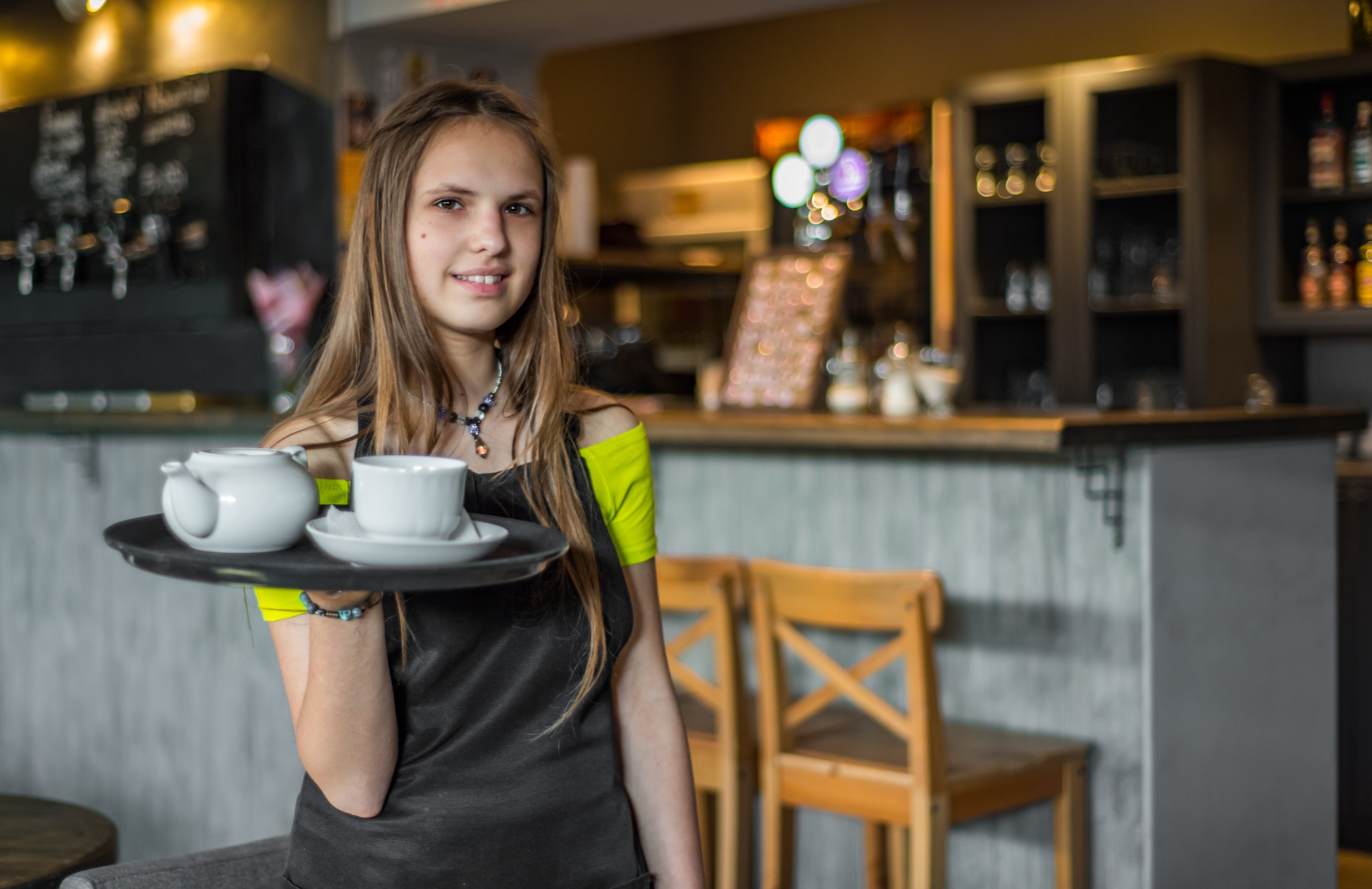Teenage girl working in a cafe carrying a tray which holds a cup and saucer and a teapot.
