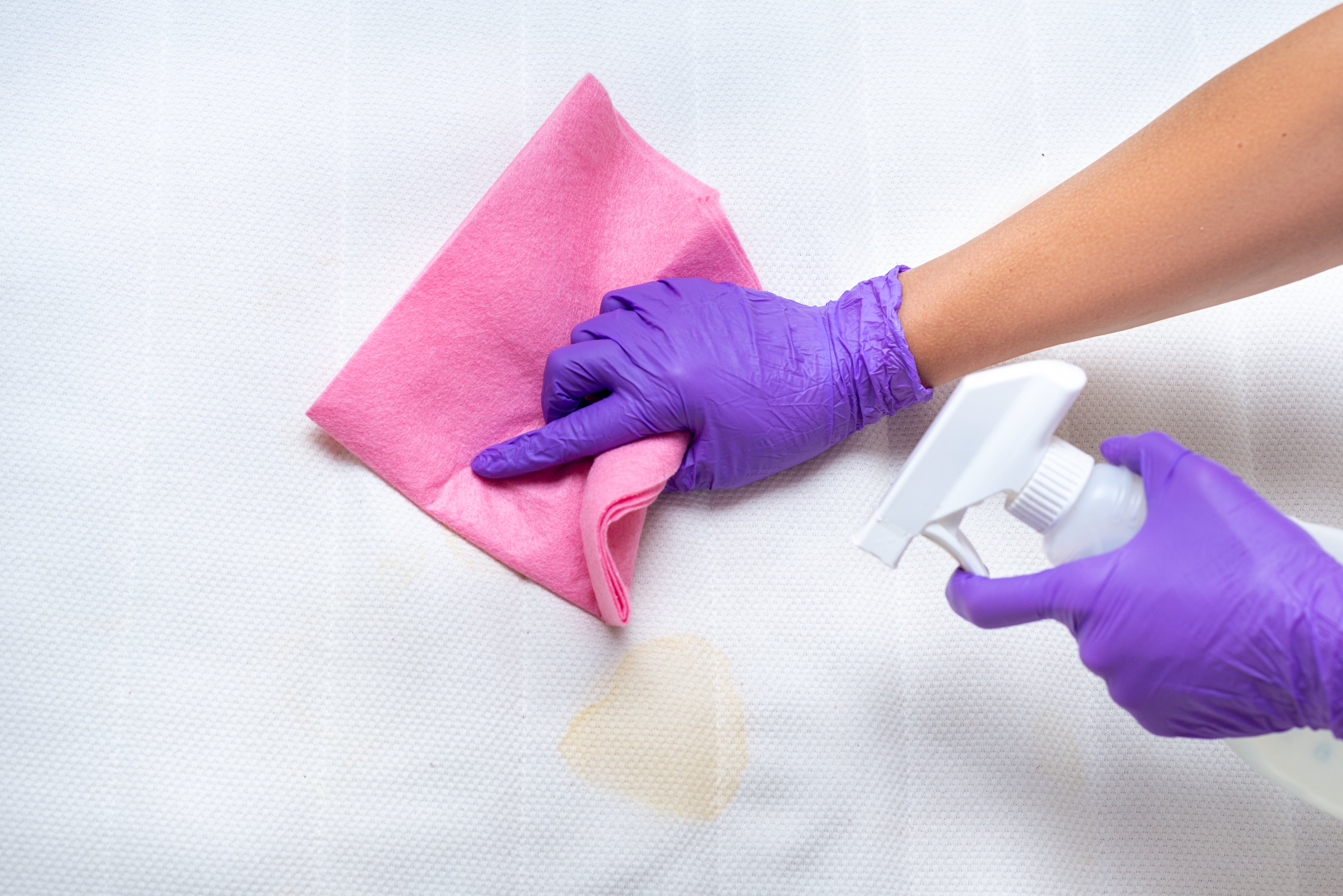 A latex gloved hand wipes a stain on a mattress with a microfibre cloth. The other latex gloved hand holds a spray bottle.