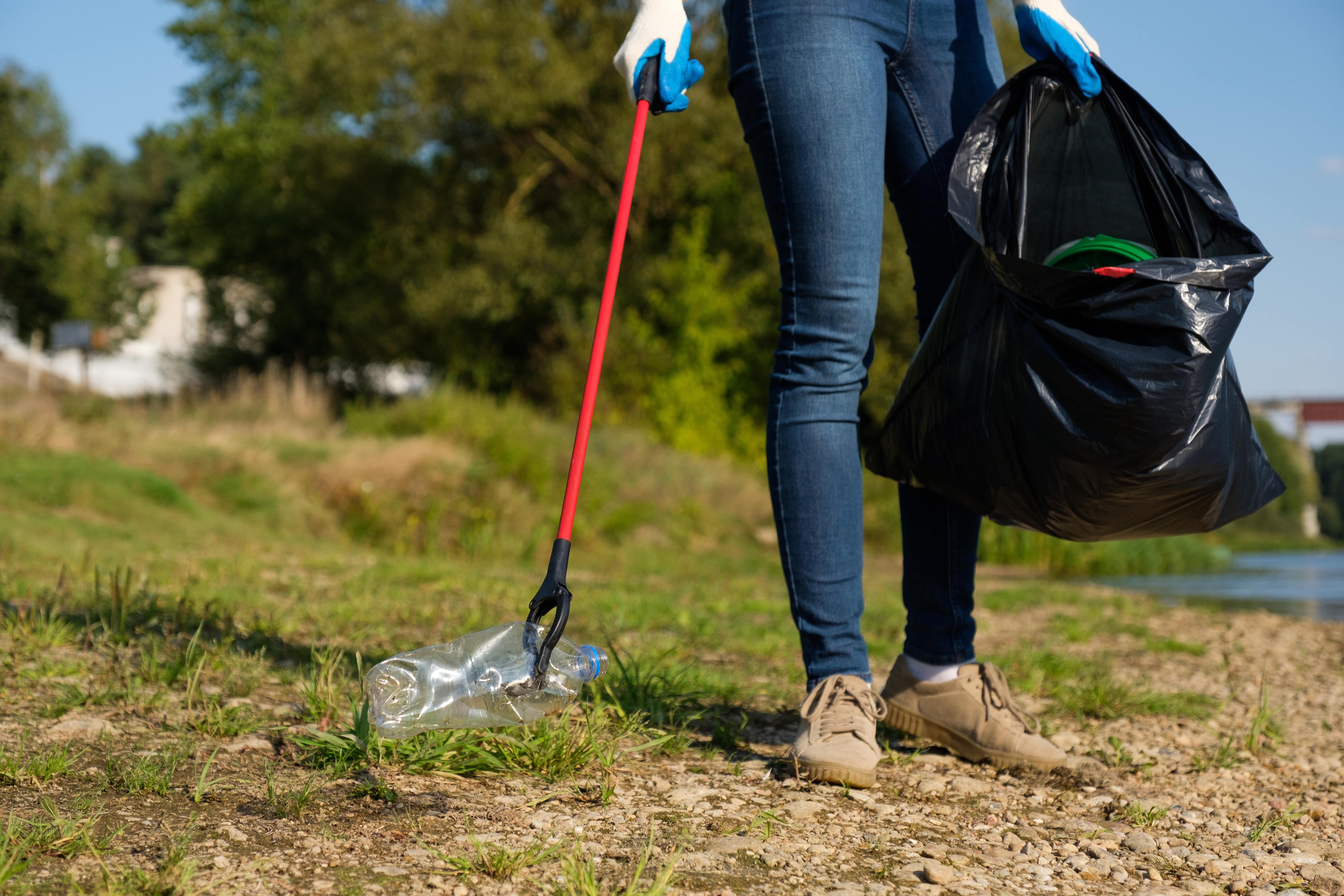 A woman with gloved hands uses a litter-picker to pick up a plastic bottle with one hand and carries an open bin bag in the other.