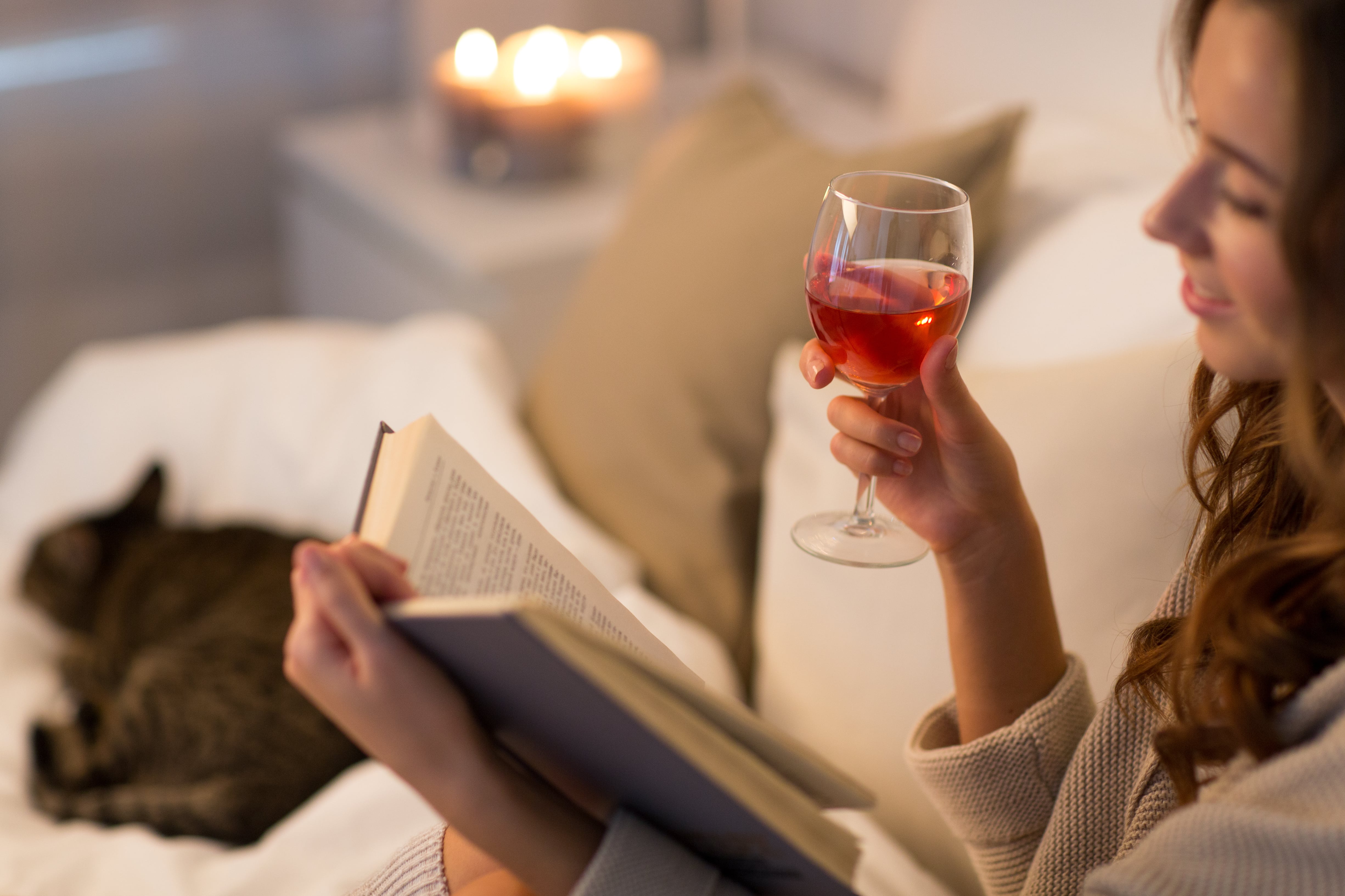 A smiling woman is relaxing, holding a book and a glass of rosé wine  A cat lies sleeping beside her and a four-wick candle glows in the background