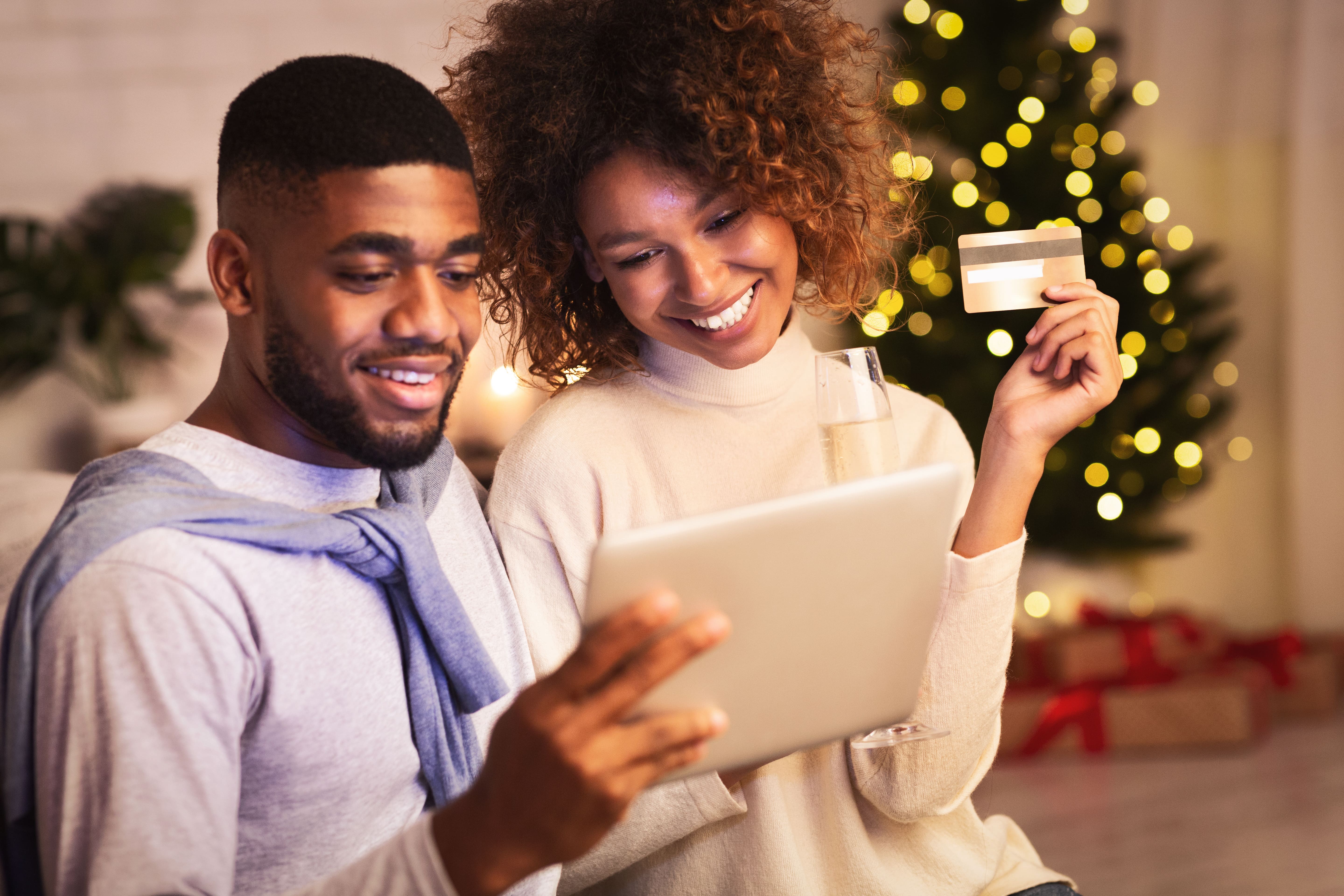 Man and woman at home smiling as they look at an electronic tablet. The woman is holding a credit card in her hand and there is a Christmas tree strewn with lights and with gifts underneath in the background.