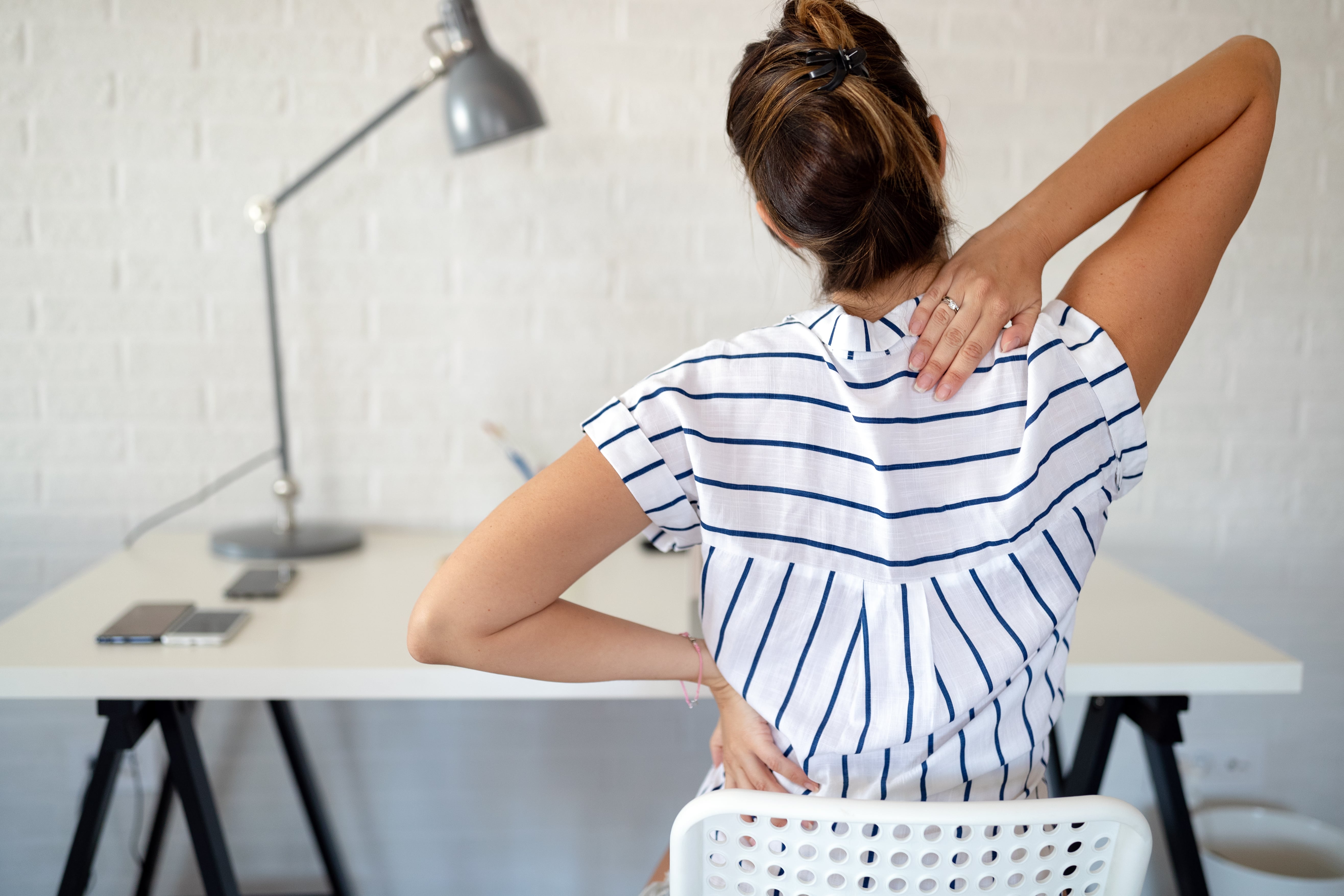 Woman at desk with her hands on her back seemingly experincing back pain