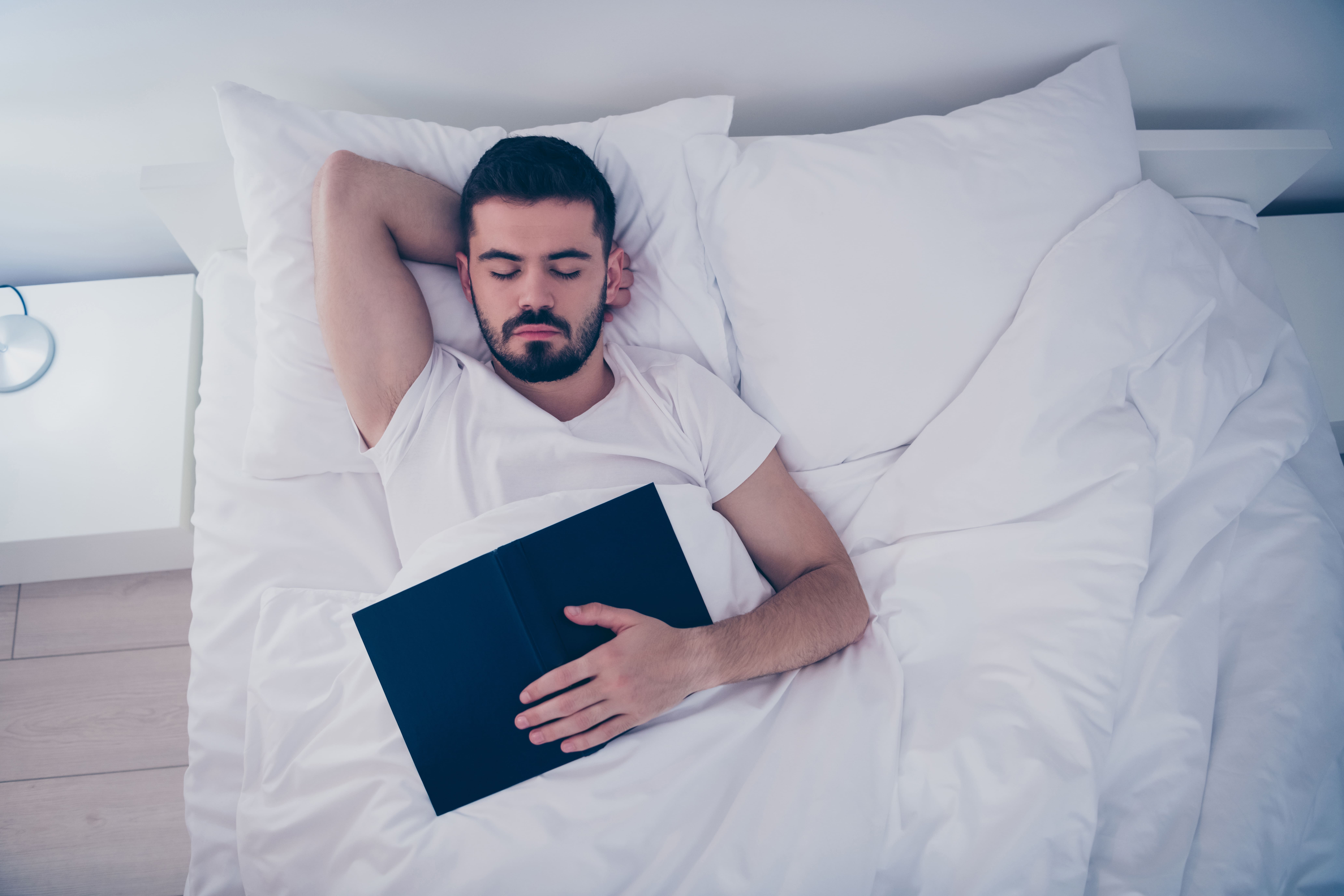 Man asleep in bed with an open book face down on his front.