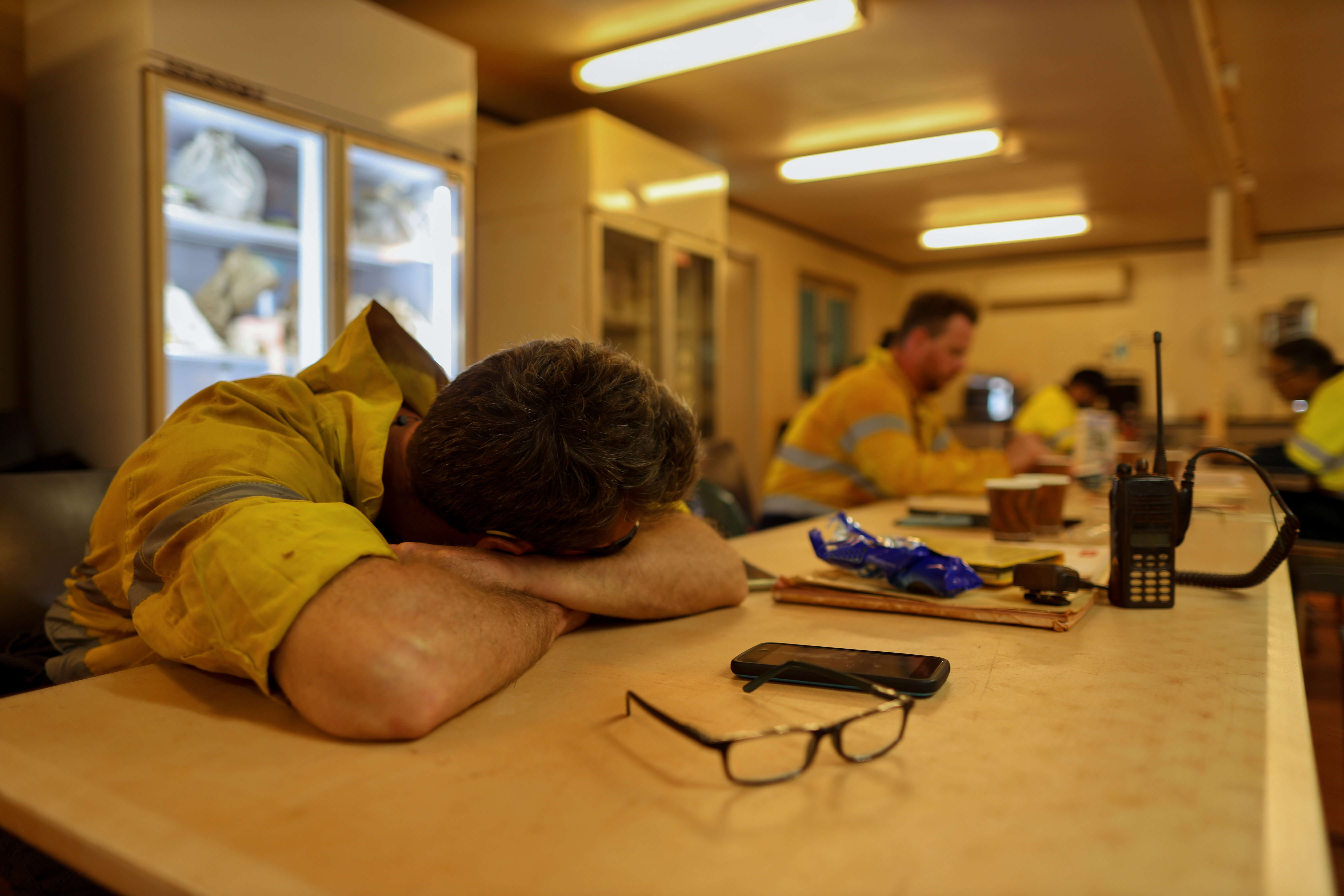 A man wearing a high visibility jacket asleep at his desk with his co-workers hard at work in the background