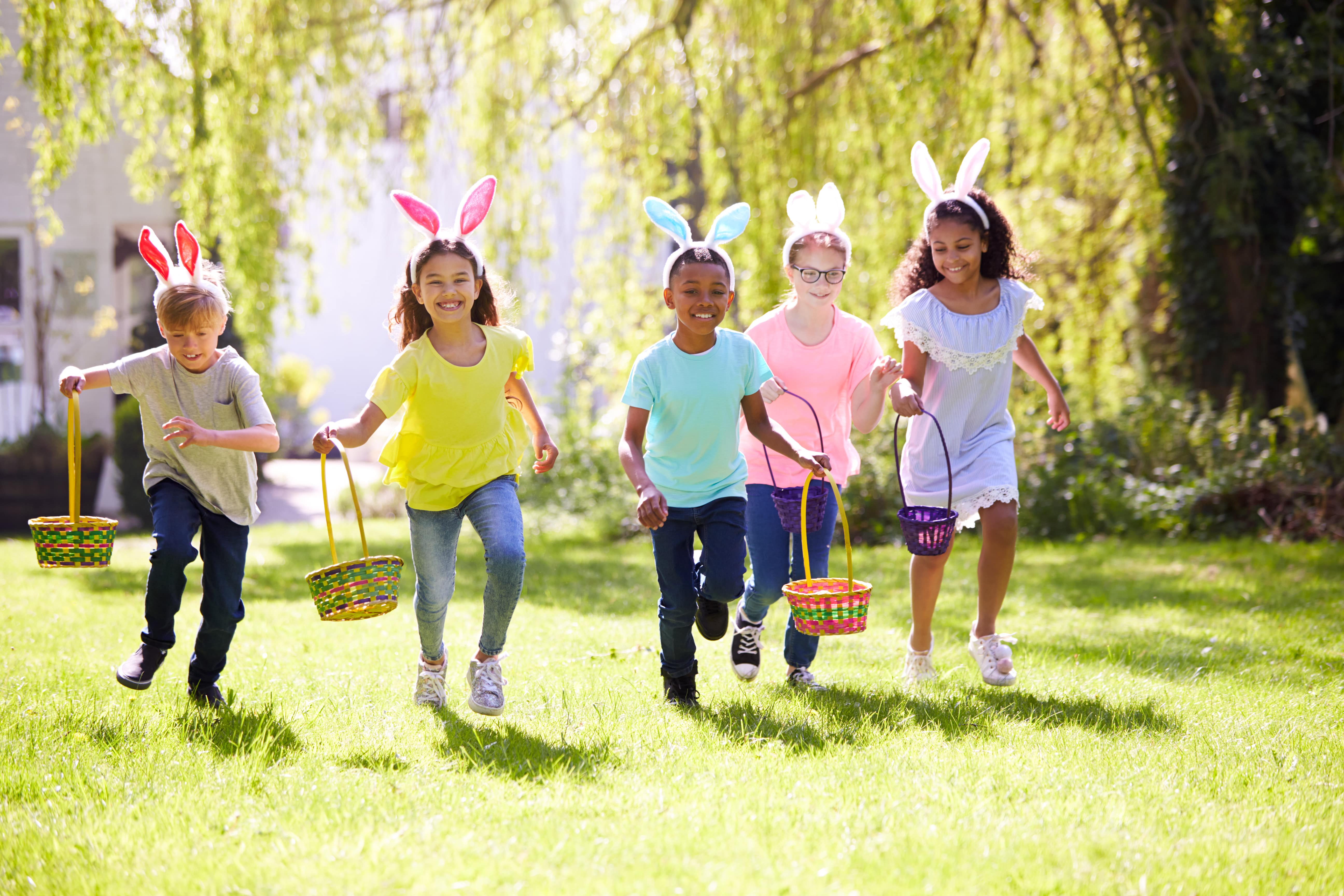 Five children wearing brightly coloured clothes and Easter bunny ears carrying Easter baskets and running across a green lawn in the sunshine.