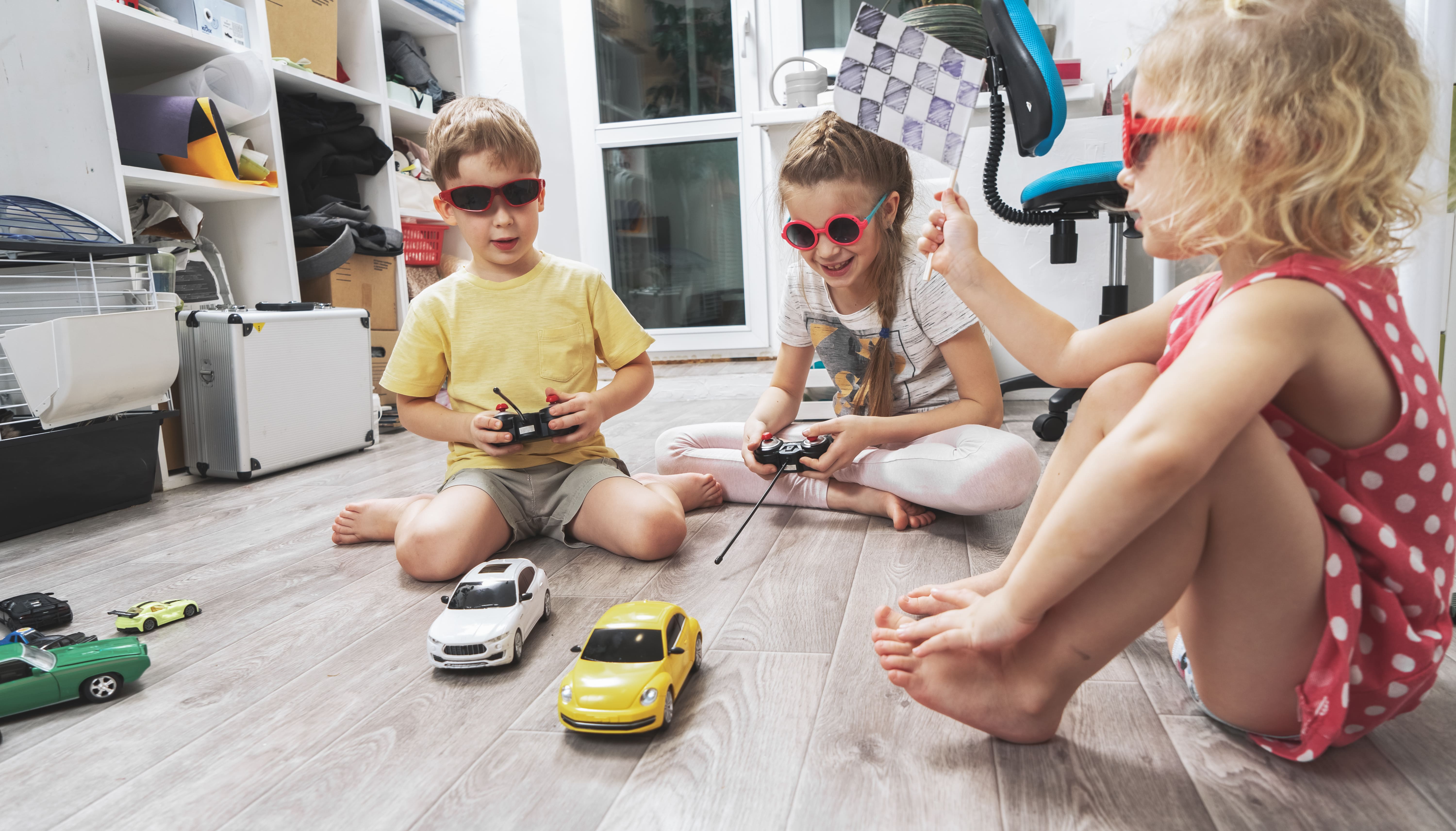 Three kids sat on the floor hosting a remote control car race while organising their toys
