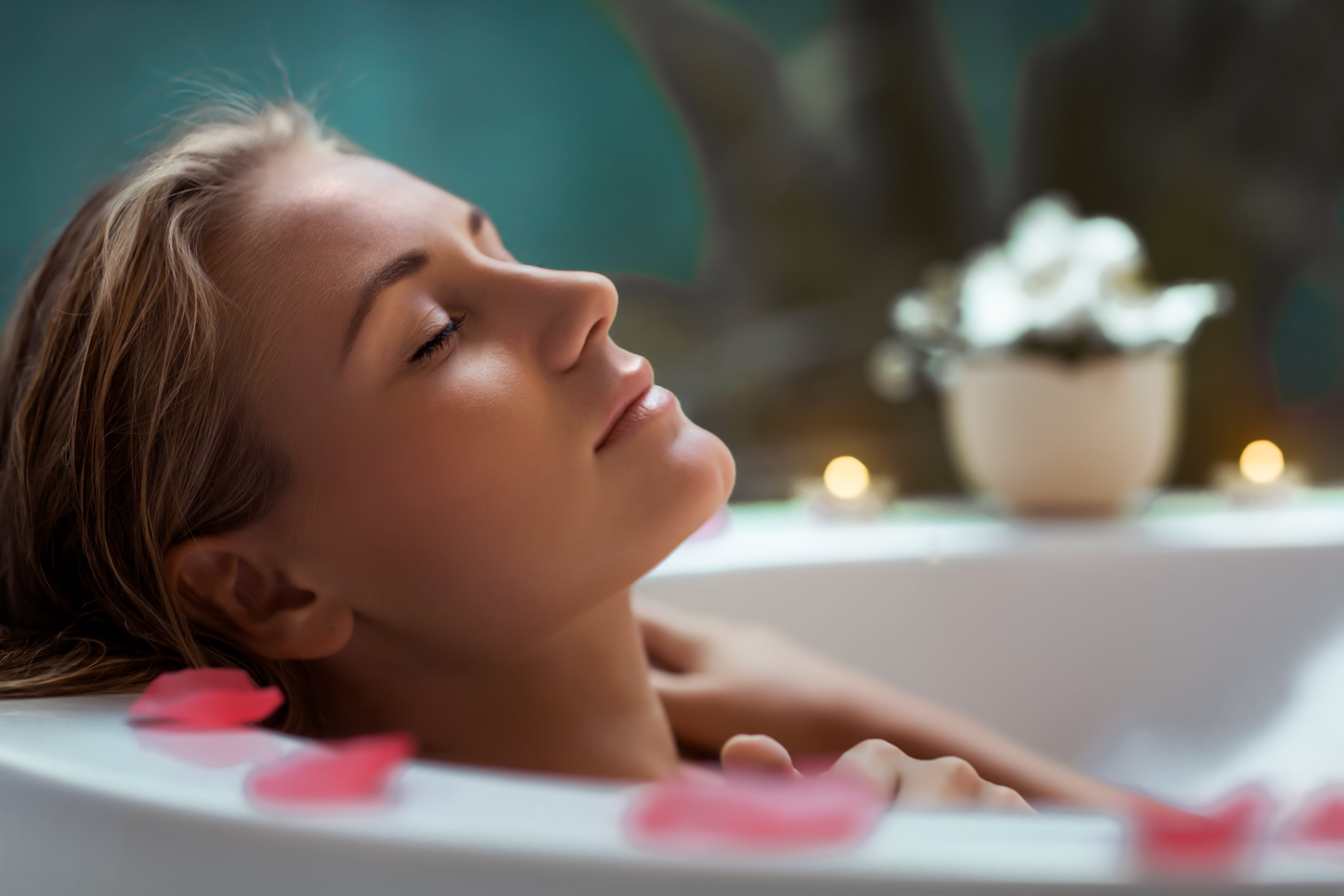 Head shot of a woman lying back in a bath with her eyes closed. There are flower petals around the rim of the bath and candles glow softly in the background.