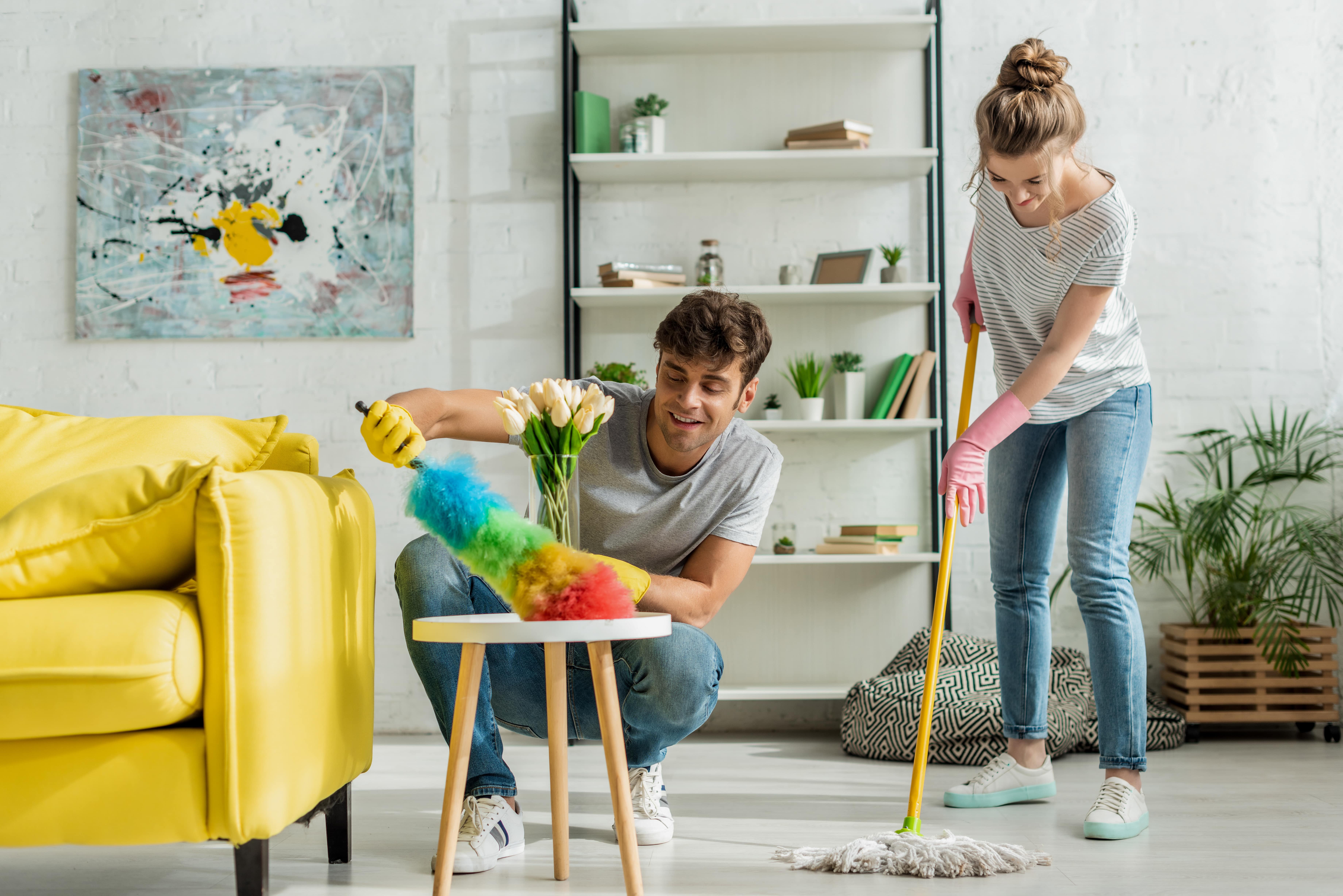 A couple working together to spring clean their home. A woman wearing pink cleaning gloves is mopping the floor while a man carefully uses a multicoloured dusting tool to remove dust for a side table.