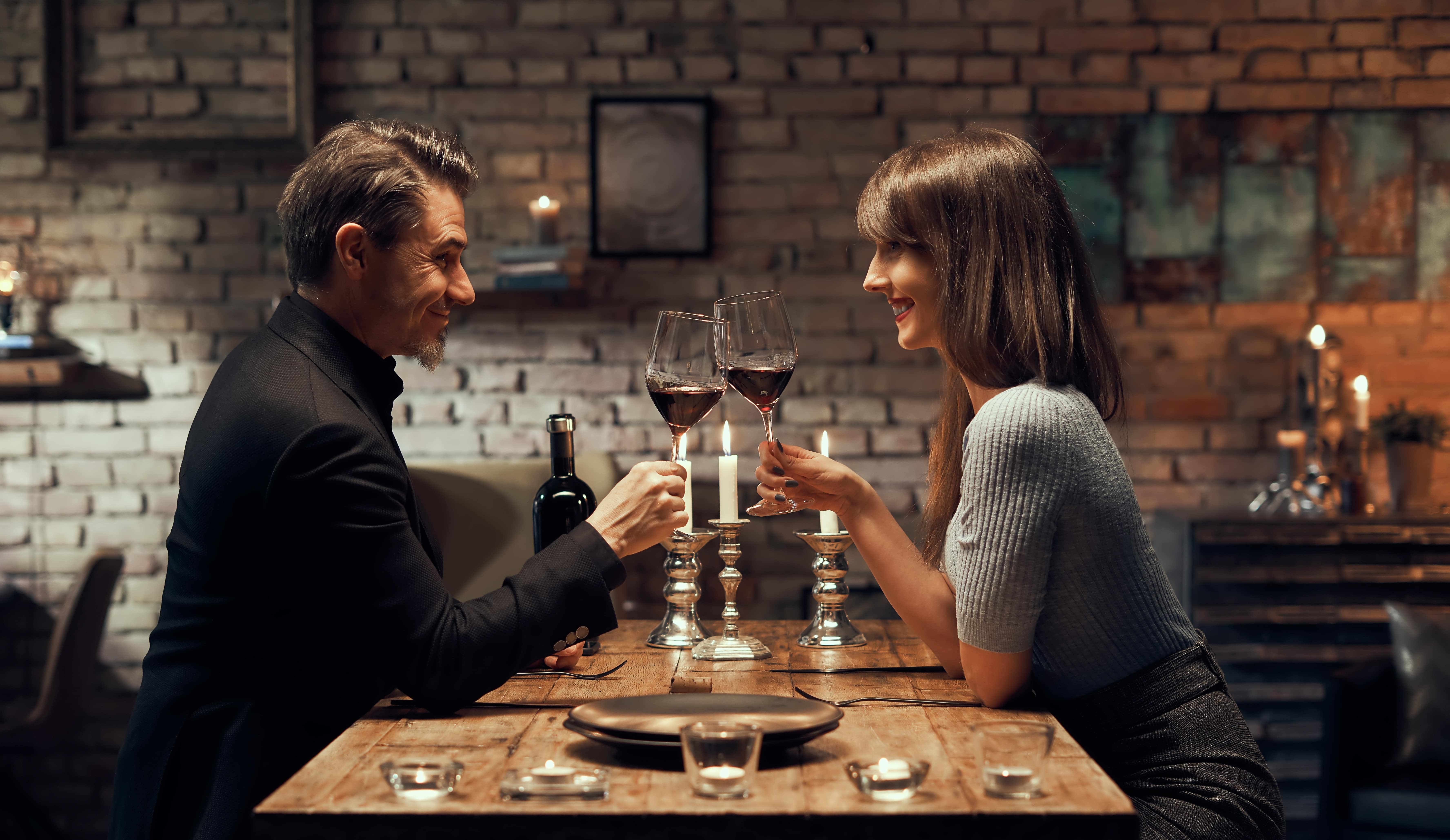A couple clinking wine glasses on a date while sat at a table in a restaurant. 