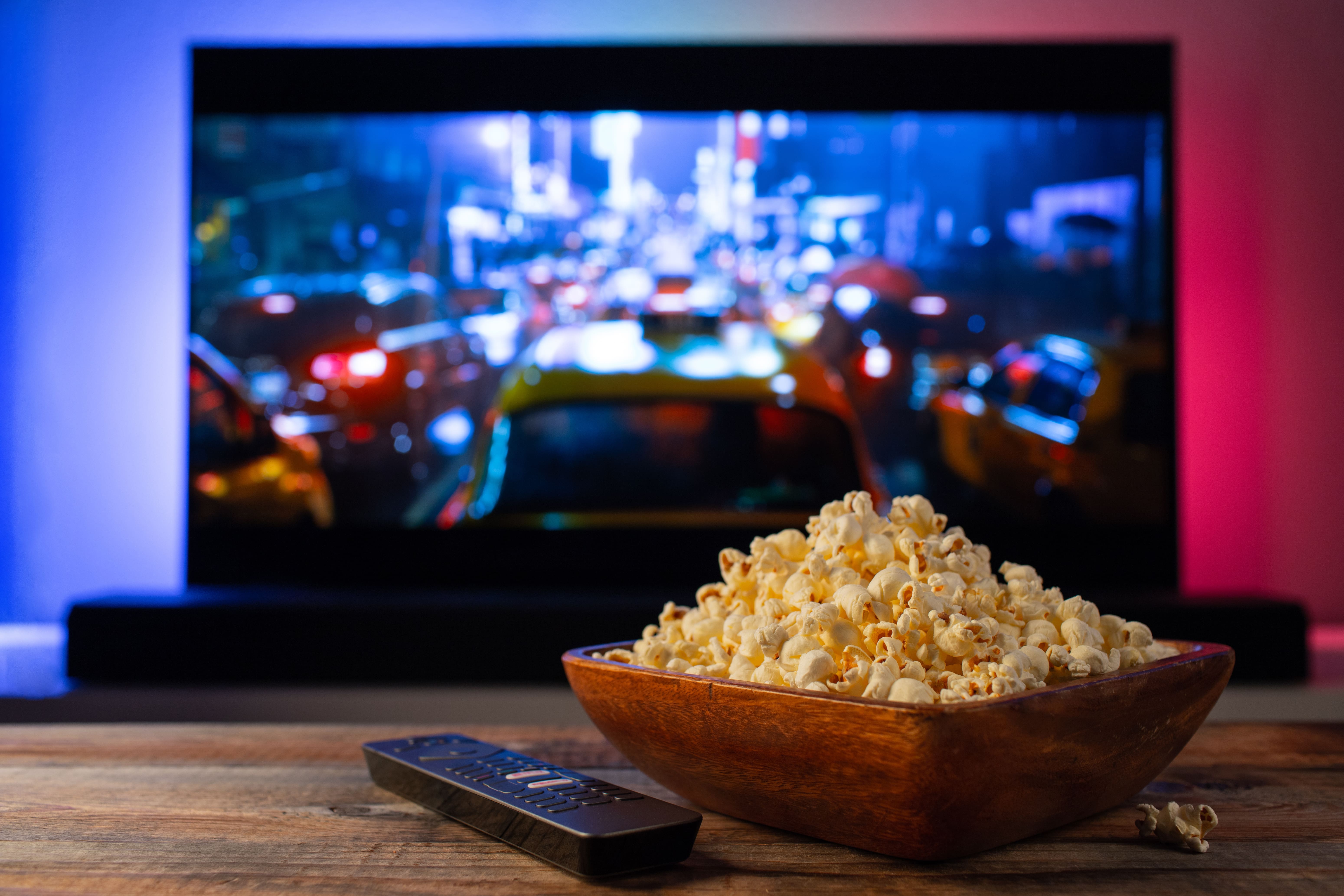 A wooden bowl overflowing with popcorn sat on a wooden coffee table next to the remote in front of a movie on a flat screen TV.