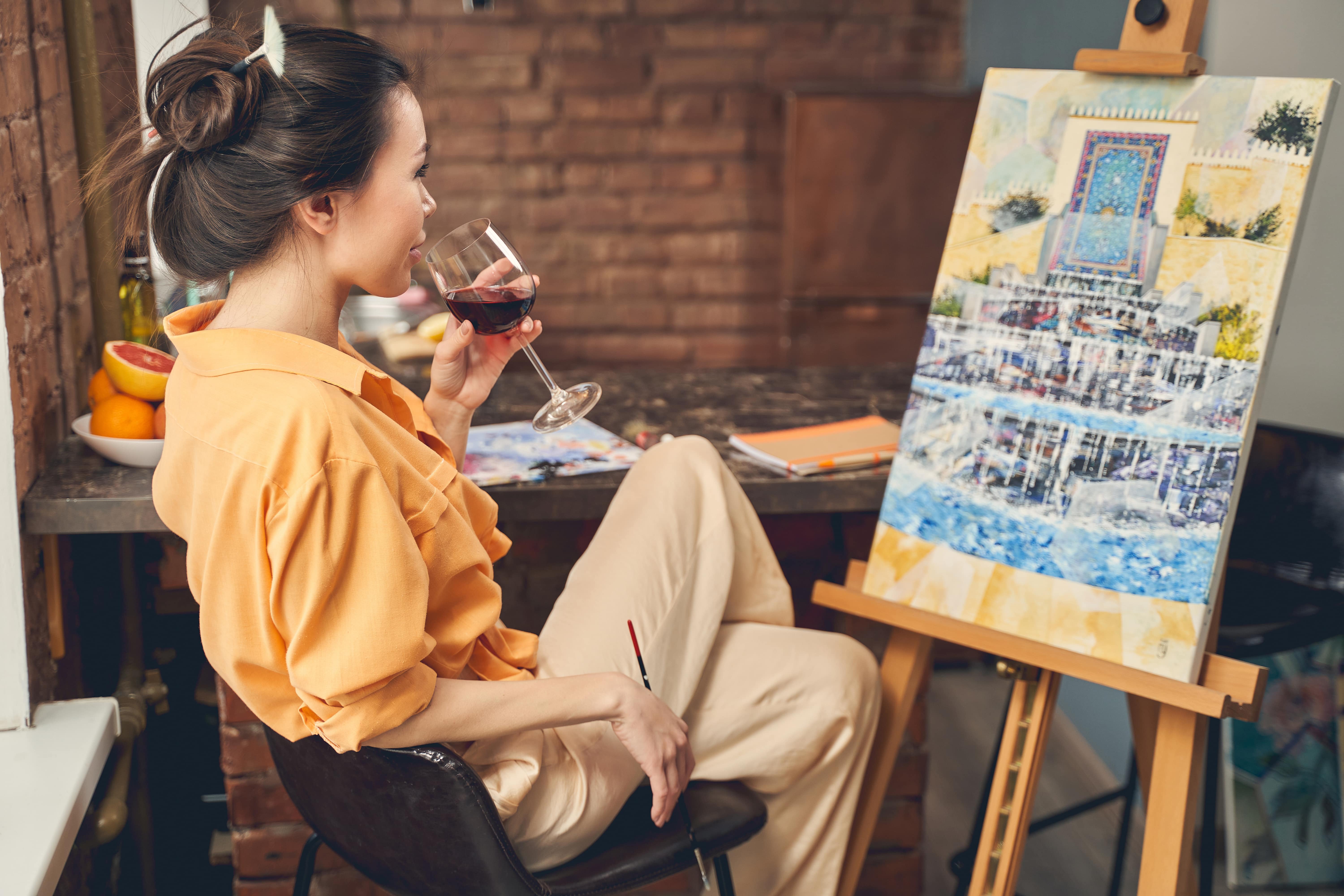 A woman sat at an easel with a glass of red wine in hand. She is about to take a sip as she surveys the progress of her masterpiece so far.