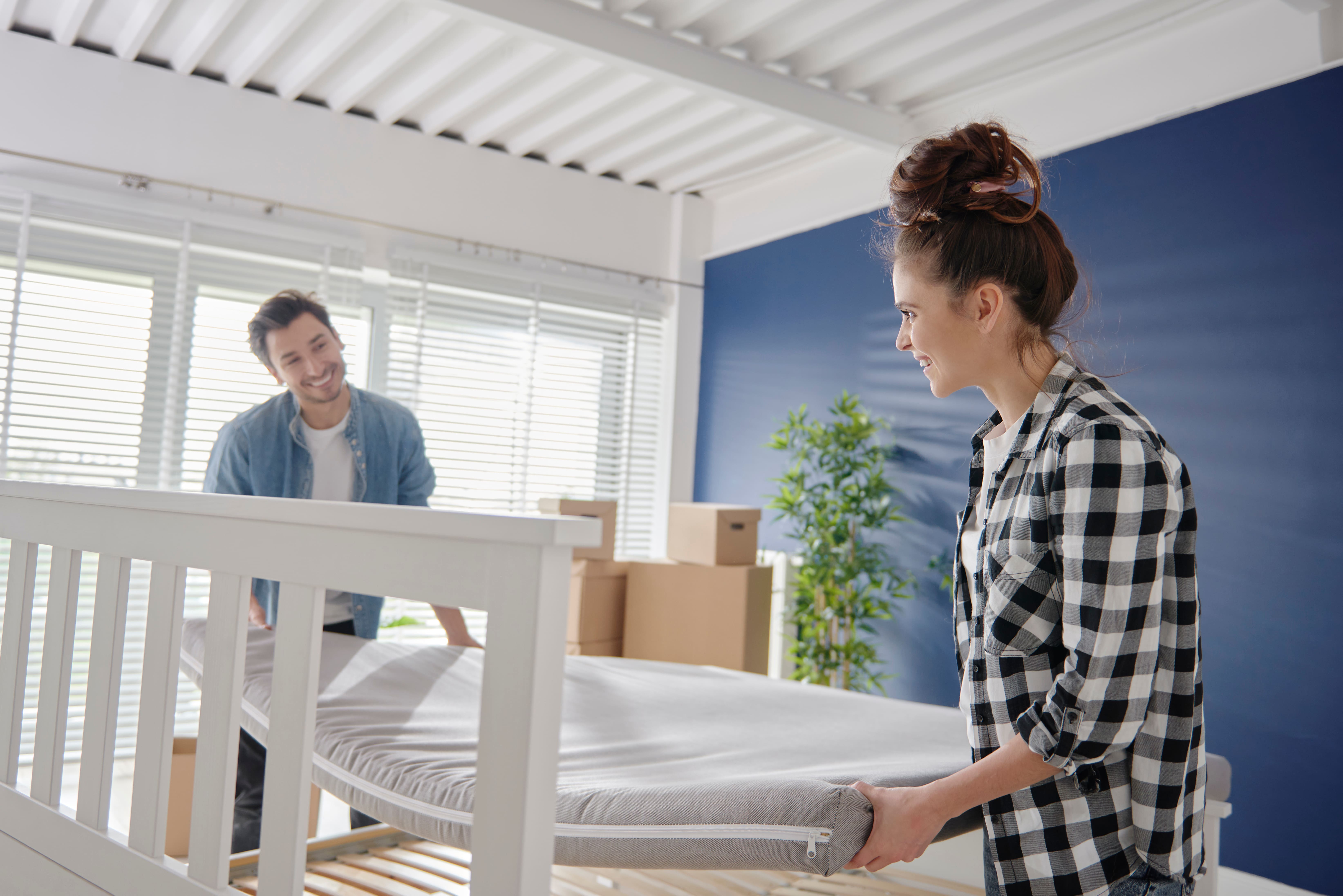 A man and woman manoeuvring their new mattress onto their new bed frame. Both are smiling and in the background, cardboard boxes and a houseplant are visible against a blue feature wall.