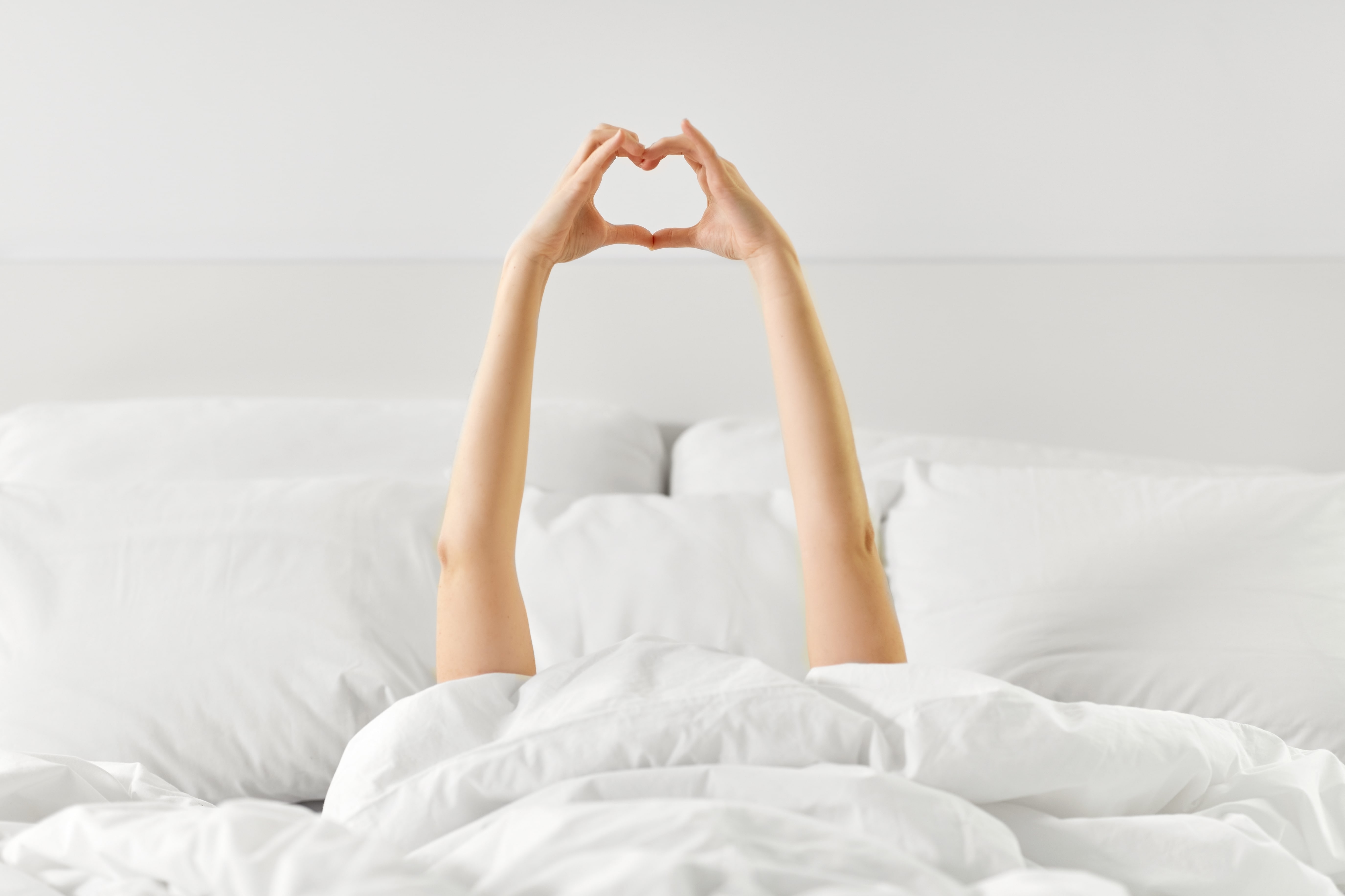 The arms of a woman in bed are outstretched with the fingers coming together to form a heart.