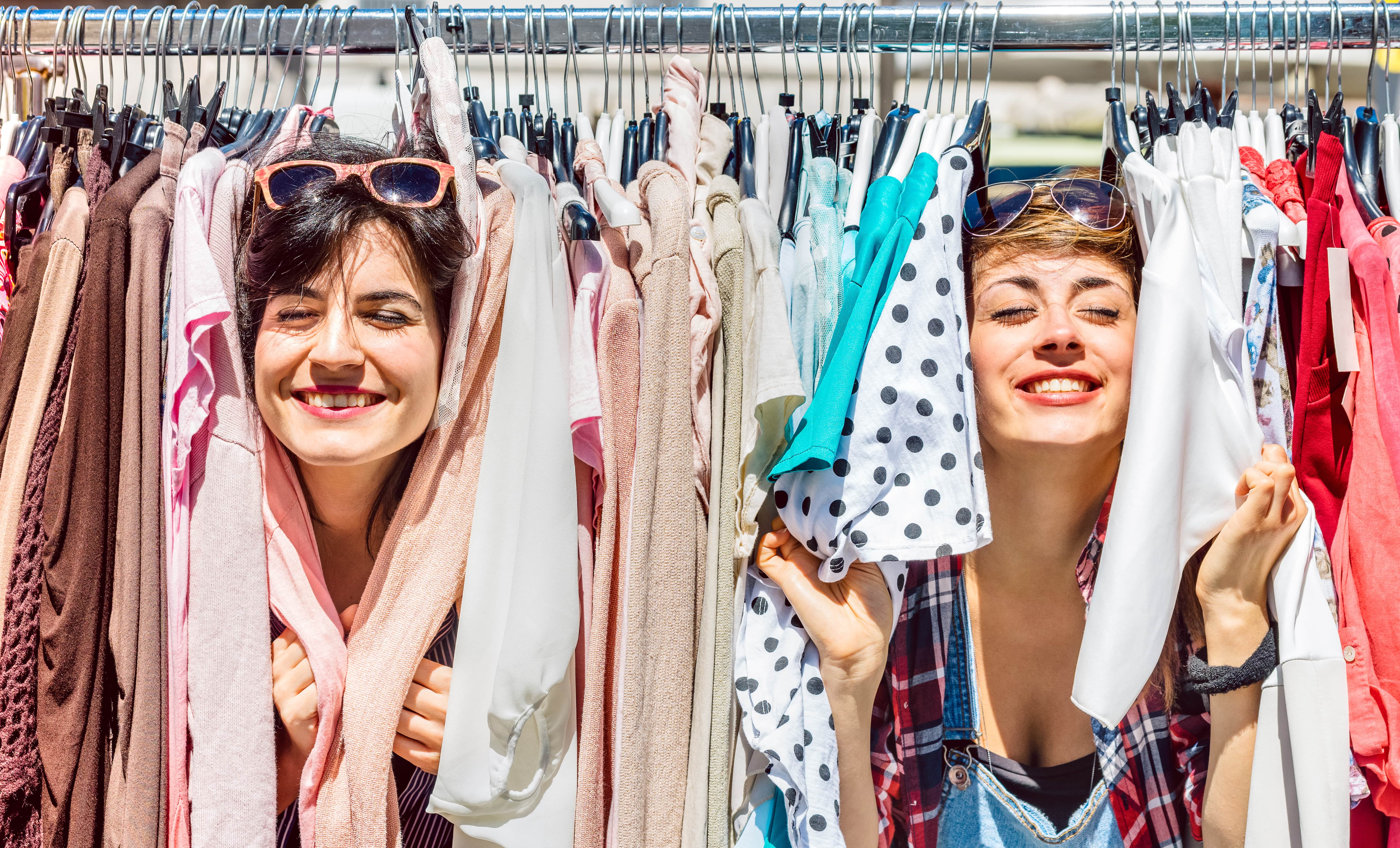 Two women with eyes closed smiling with their heads peeking through clothes on a rack.