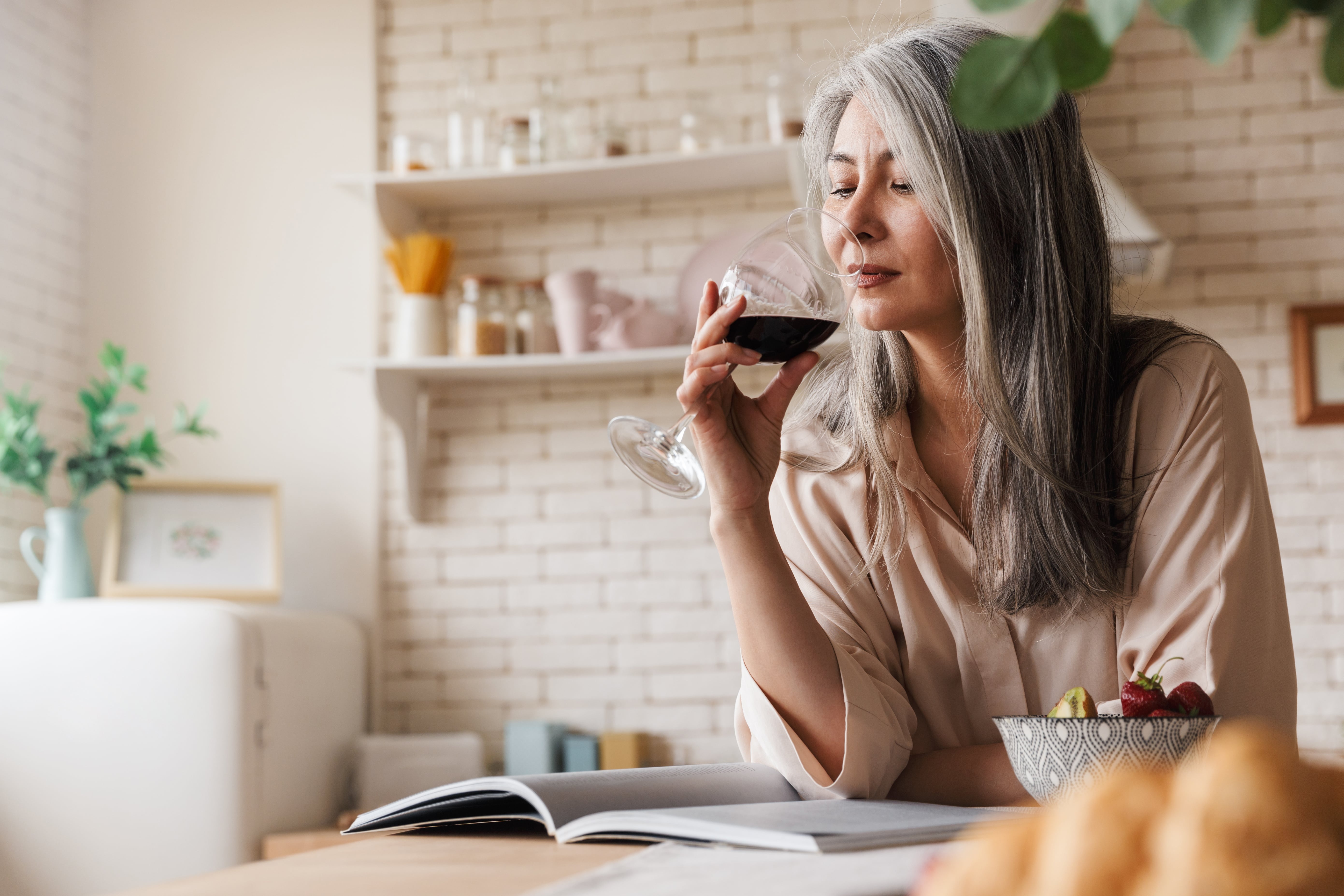 A lady within the age range for menopause drinking a glass of wine while reading an open book in a kitchen