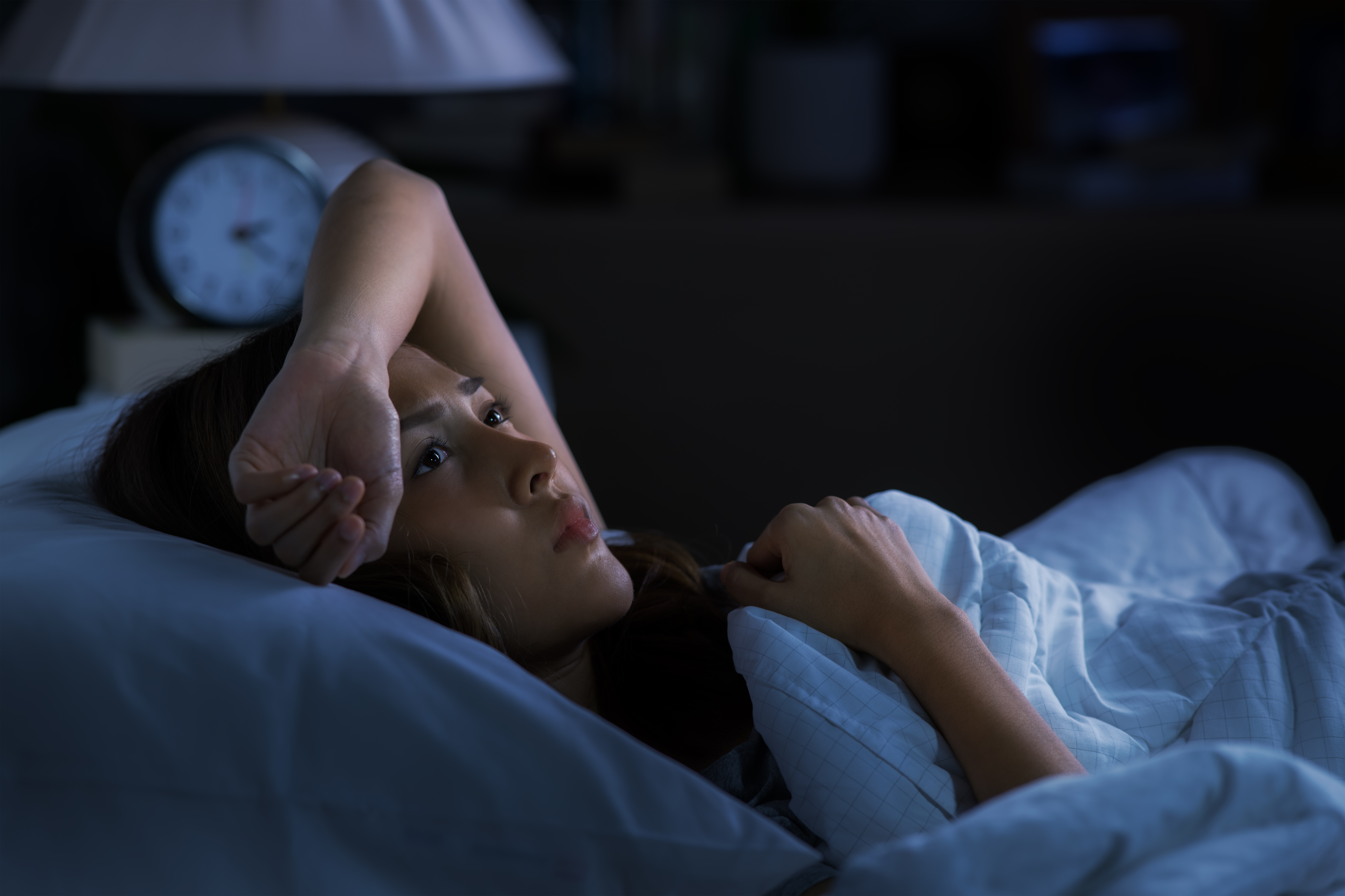 Woman in bed at night time looking stressed with one arm over her forehead.