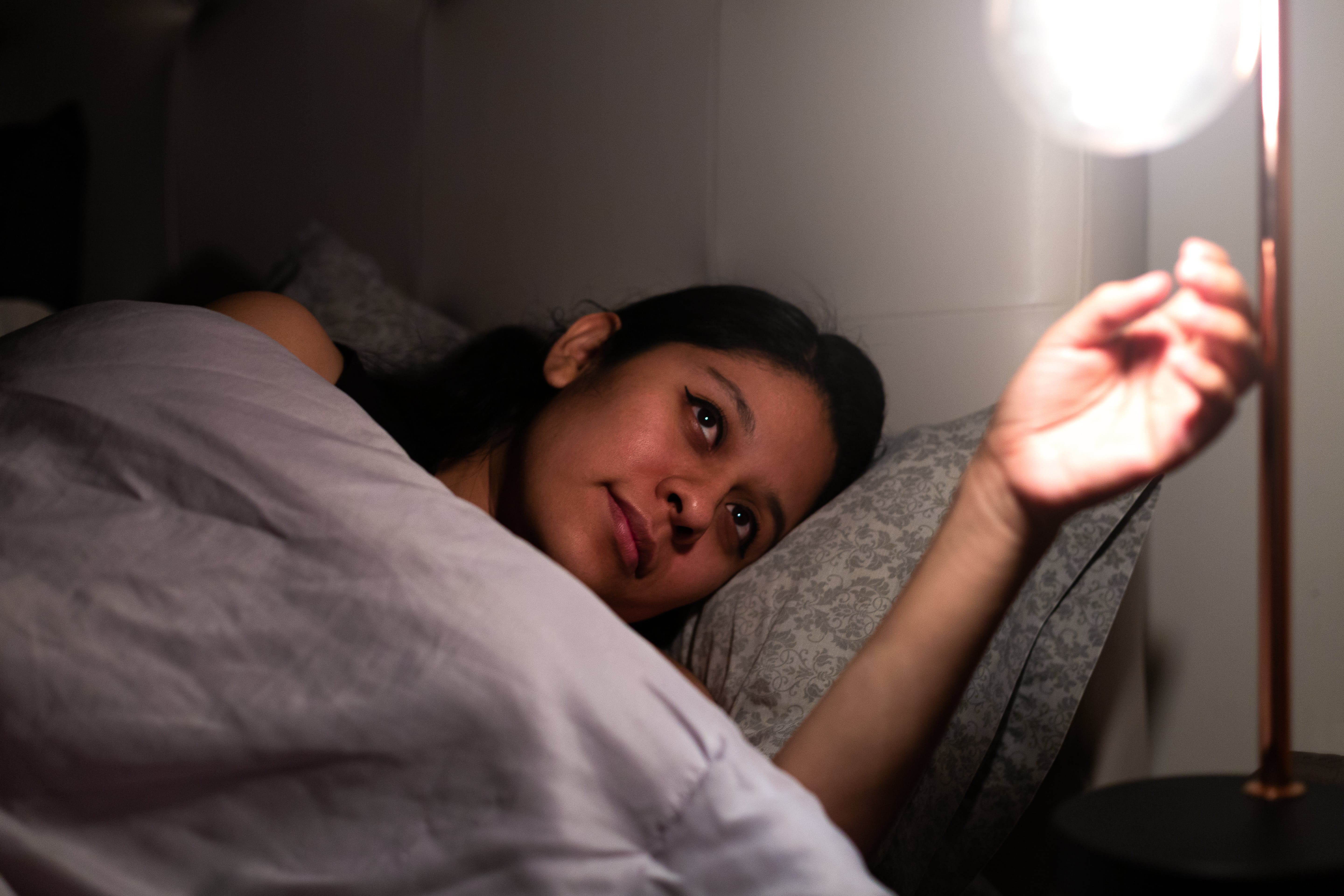 Woman in bed reaching to turn off the lamp light at the side of her bed.