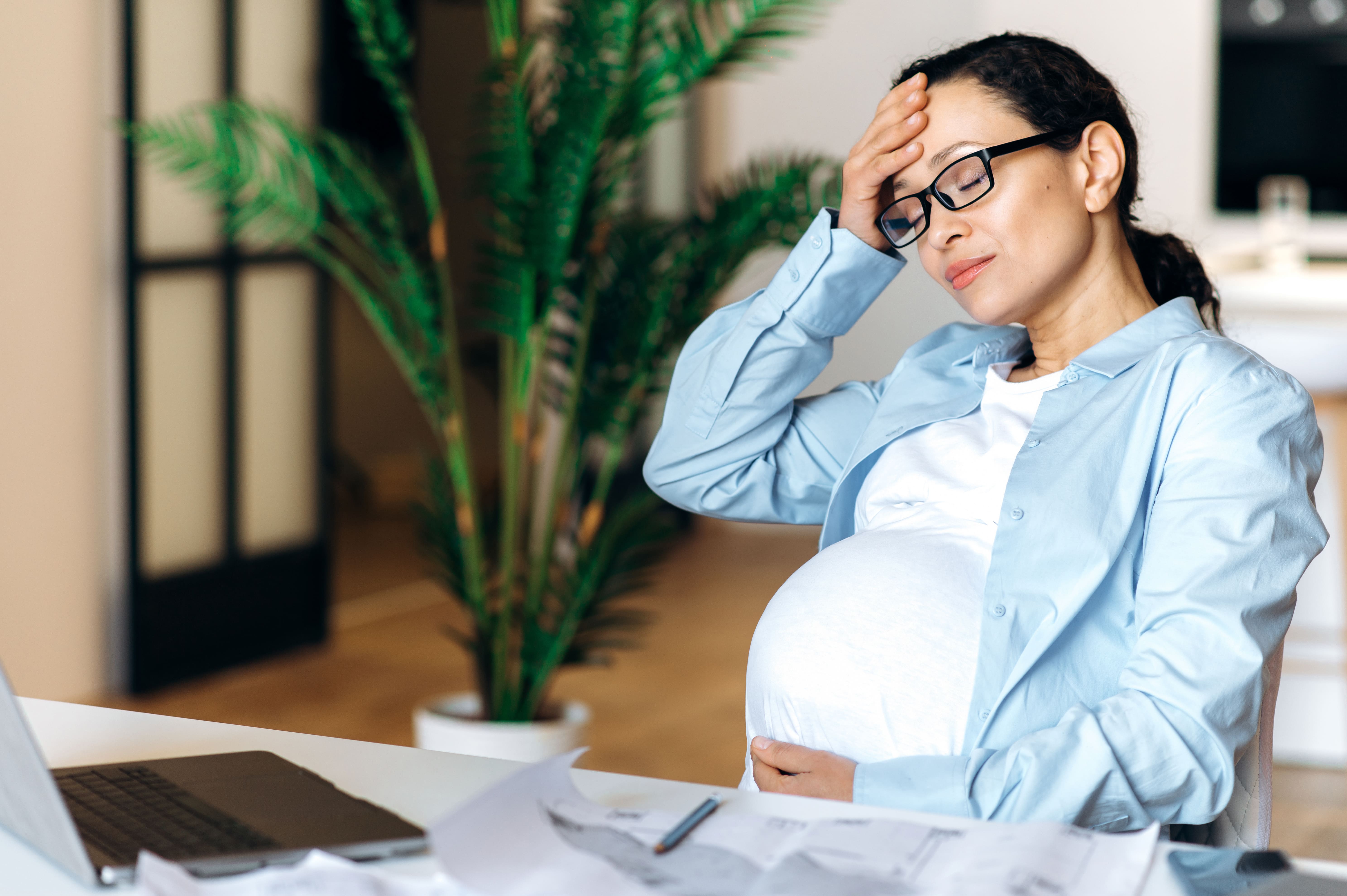 Woman at desk with eyes closed with one hand to her head and the other resting on her large pregnancy bump