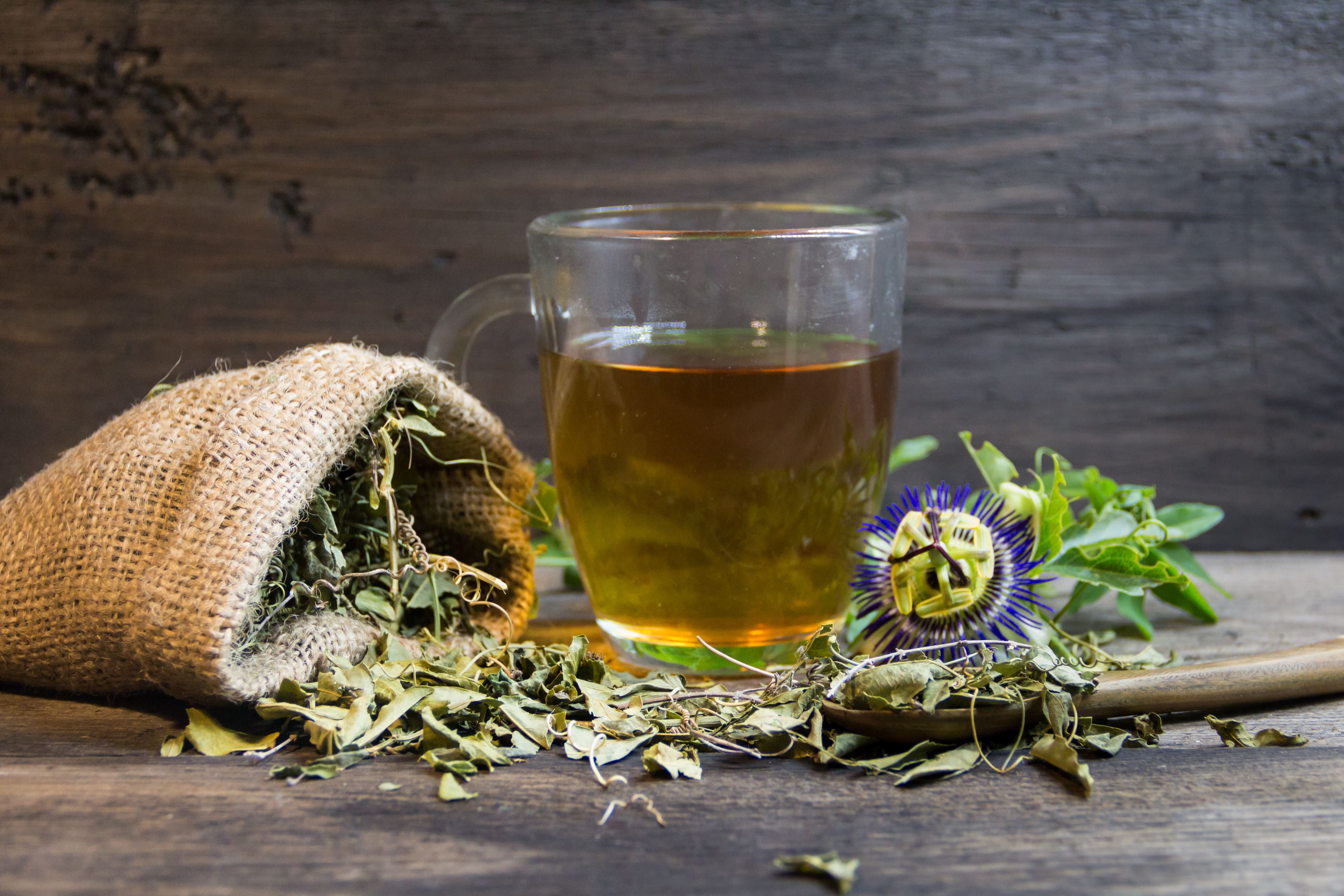A glass mug of tea made using passion flower extract. A hessian sack filled with dried passion flower lies on one sider and a passion flower in its original, fresh form lies on the other 