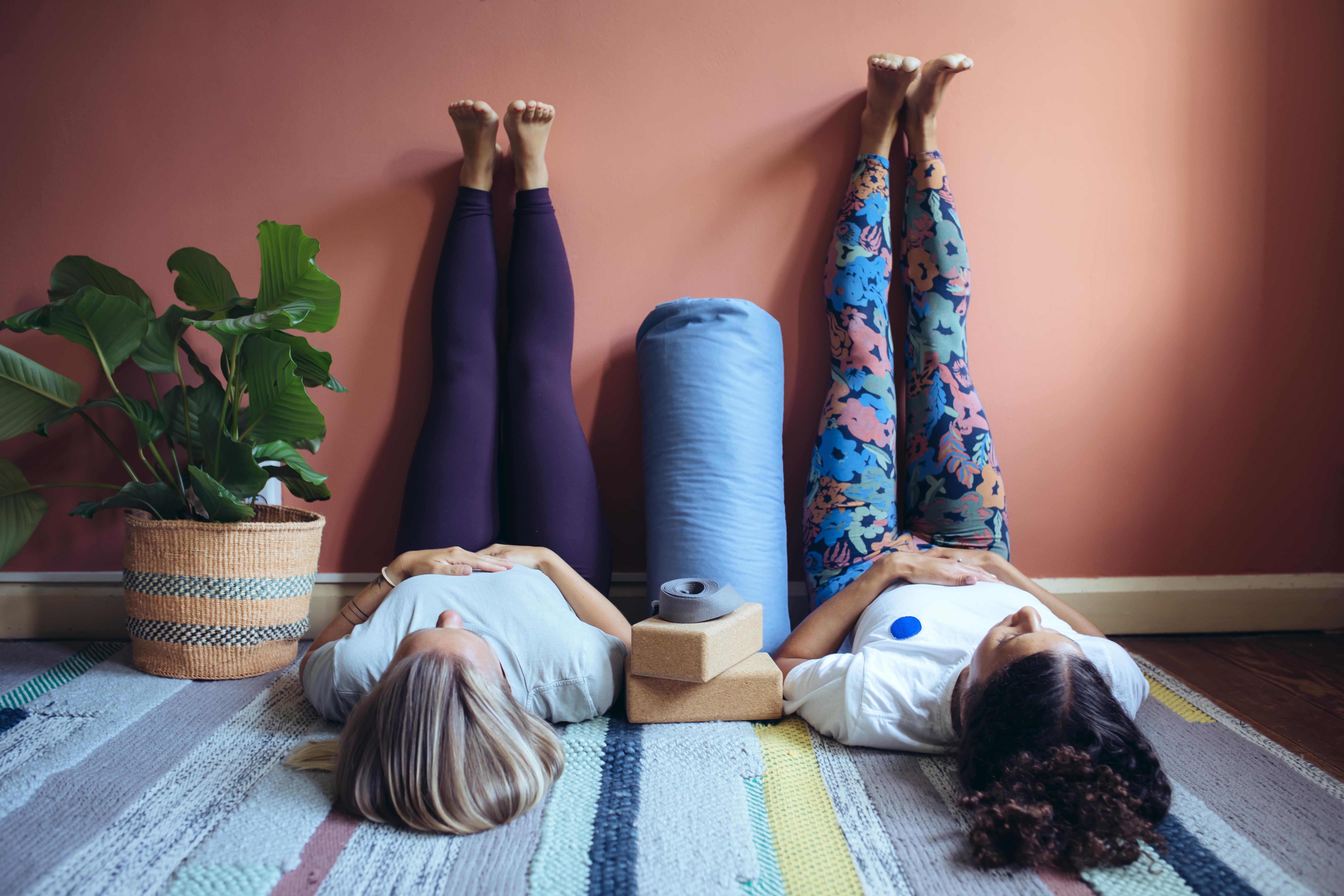 Two ladies practicing the legs up the wall yoga pose side by side. The wall is a burnt orange colour and the ladies are lying on a multicoloured striped rug